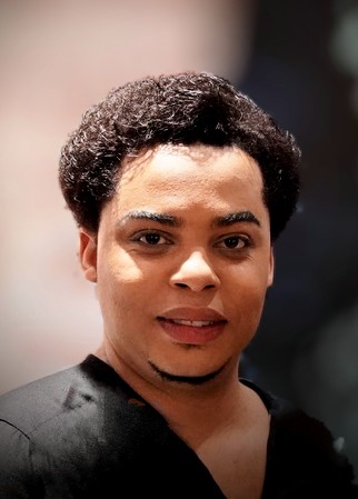 Portrait of a person with short curly hair, smiling, wearing a dark top, with a blurred background.