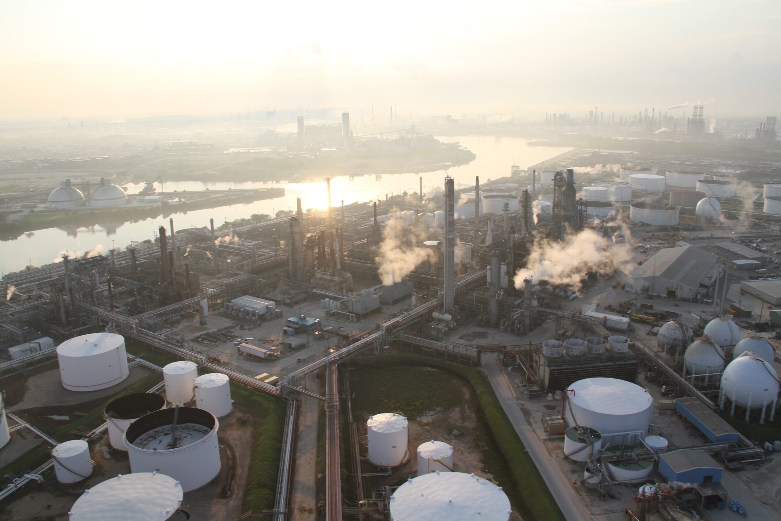 Industrial plant with storage tanks and chimneys emitting smoke, near a river with a city skyline in the background.