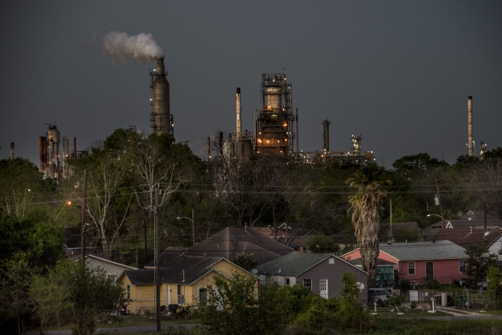 Industrial factory with smokestacks emitting smoke over residential neighborhood with houses and trees.