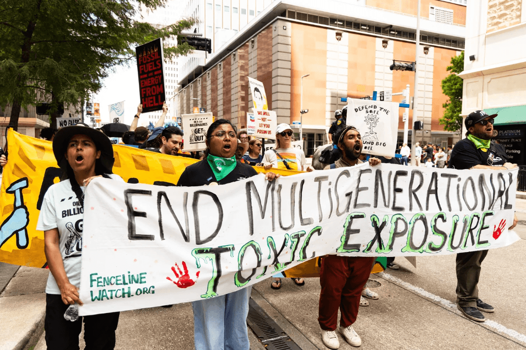 Group of people holding a protest banner on a city street, with signs and trees in the background.