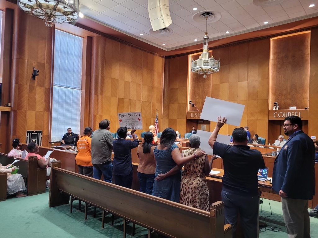 People standing and raising hands in a room with wood-paneled walls and a chandelier, possibly in a meeting or protest.