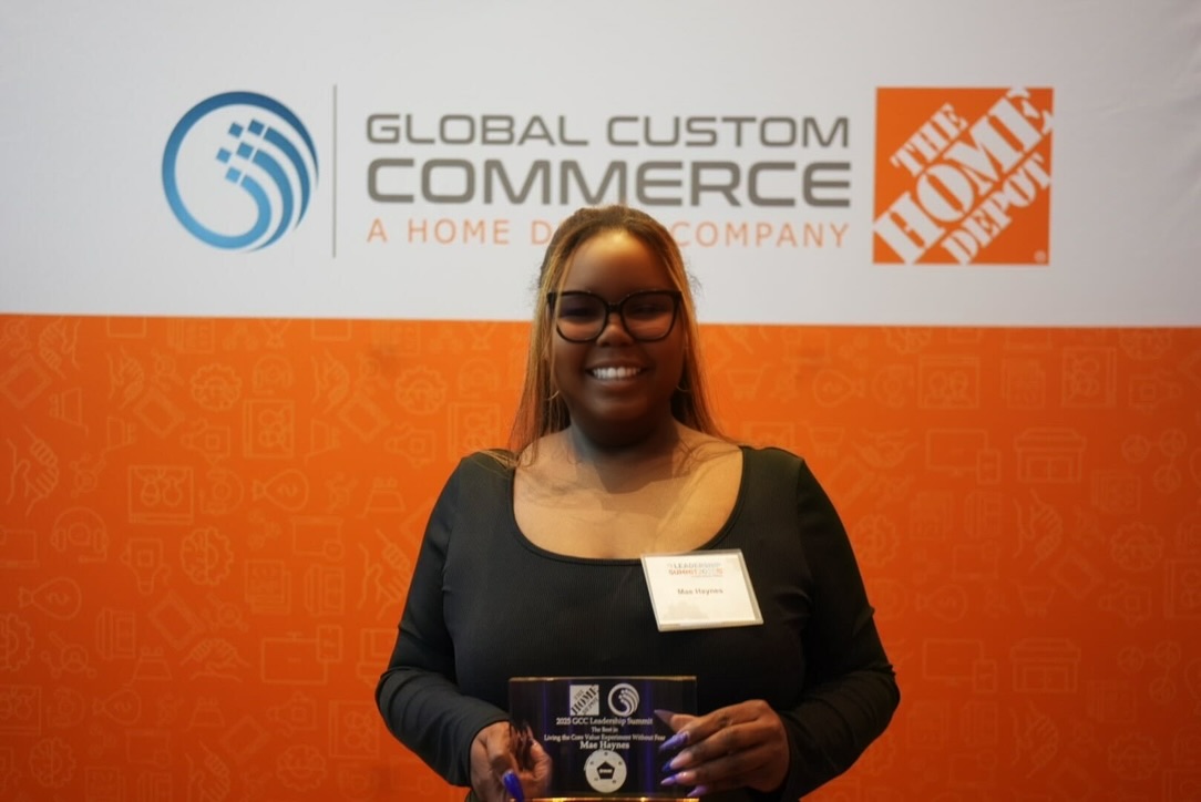 Smiling woman holding an award in front of a backdrop with logos, wearing glasses and a black top.