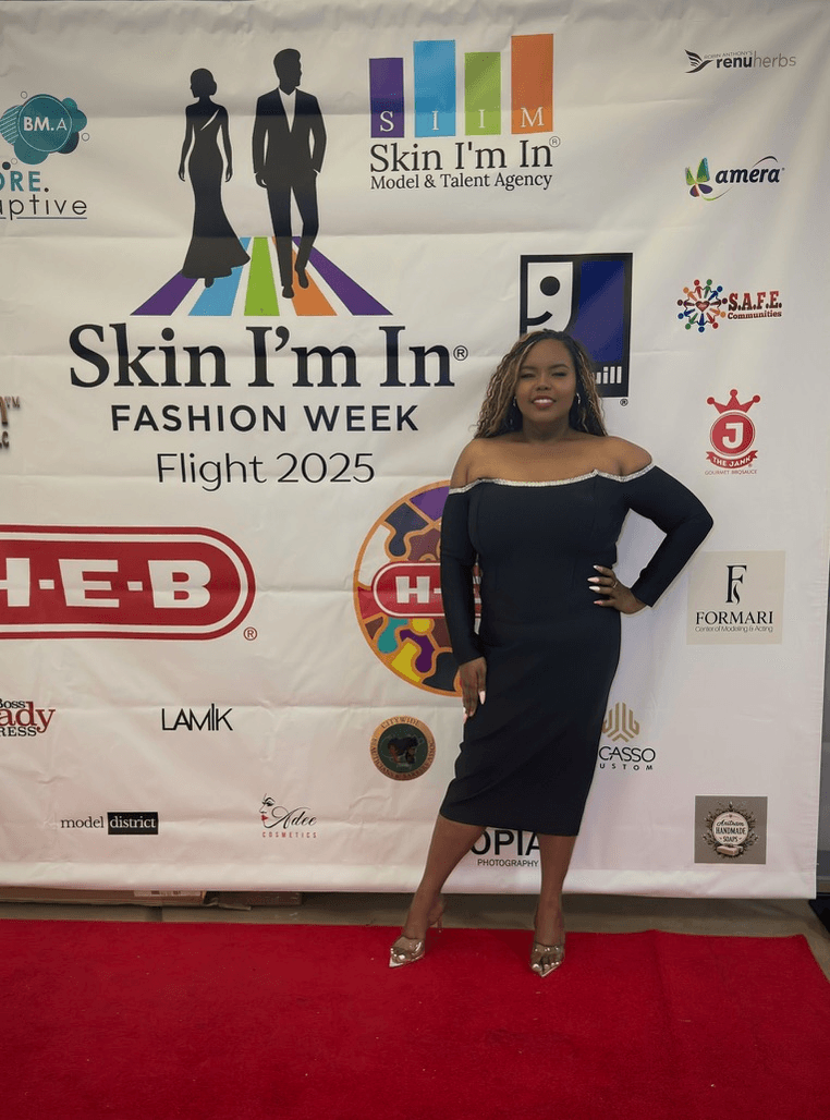 Woman in black dress standing on red carpet in front of a promotional backdrop for Skin I'm In Fashion Week Flight 2025.