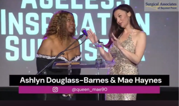 Two women on stage, one speaking into a microphone, the other holding a trophy, with a large screen behind them.