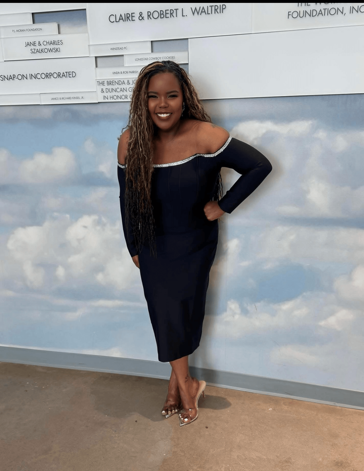Woman in a black dress with off-shoulder design, smiling, standing against a sky-themed wall, hands on hips, heels, long hair.