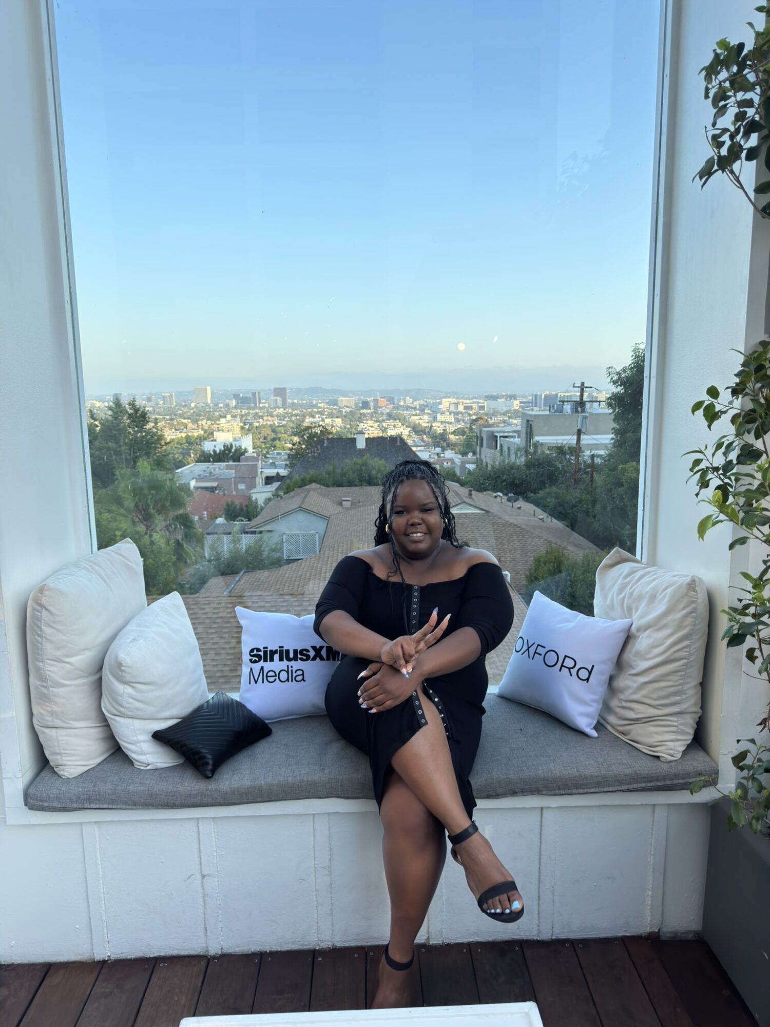 Woman sitting on a window seat with pillows, smiling, with a city view and blue sky in the background.