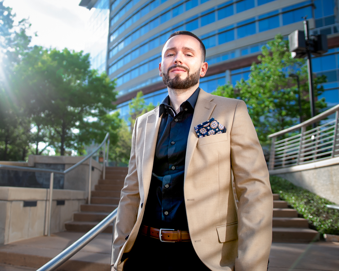 Man in beige suit and black shirt standing outdoors near stairs and modern building, smiling with sunlight behind him.