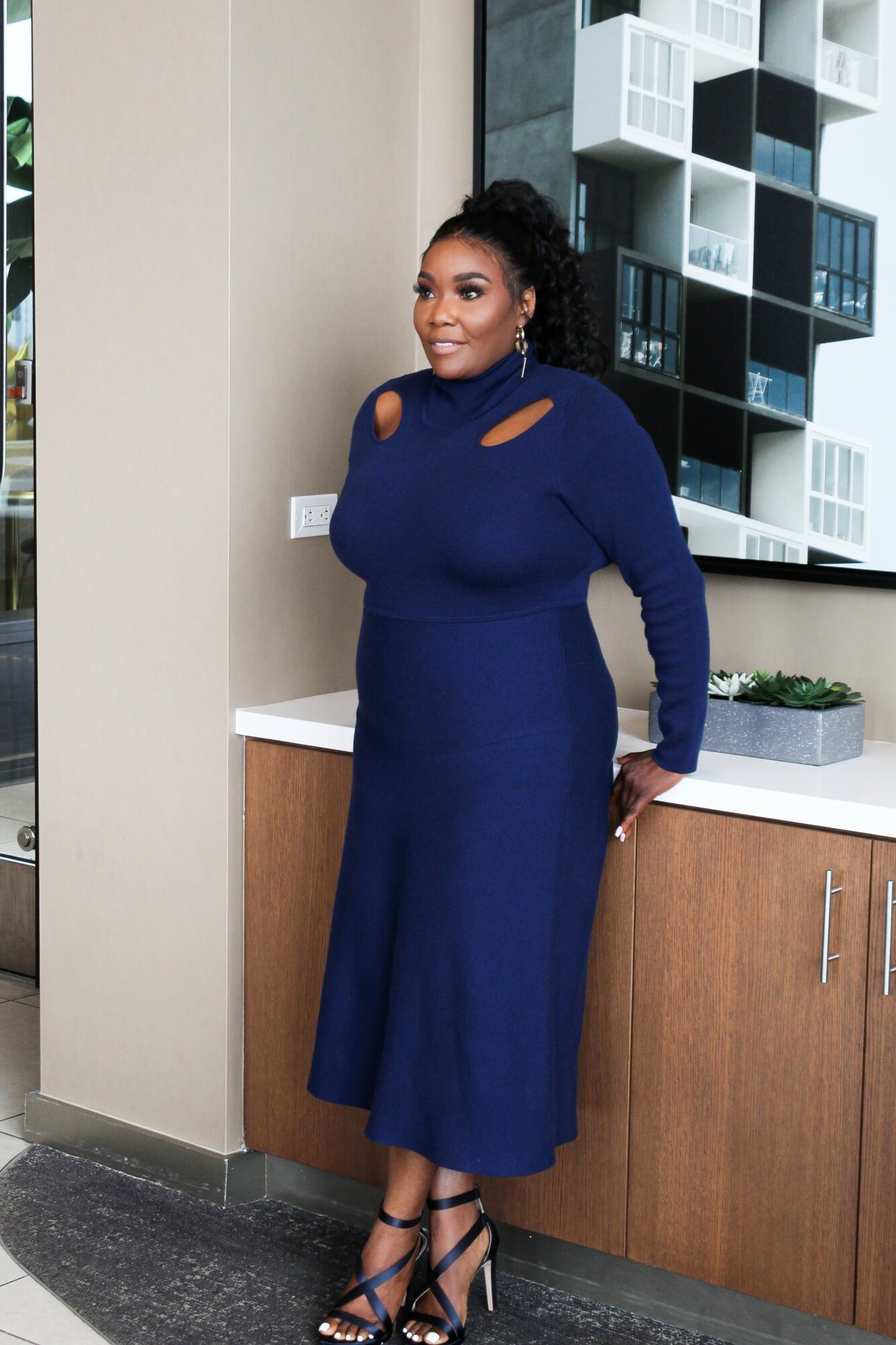 Woman in a long blue dress with cutouts on shoulders, standing indoors near a kitchen counter, smiling, wearing high heels.