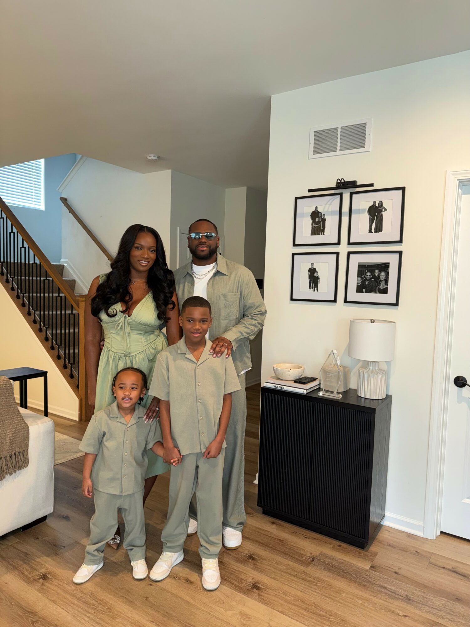 Family of four standing in living room, smiling, with staircase and framed pictures on wall behind them.