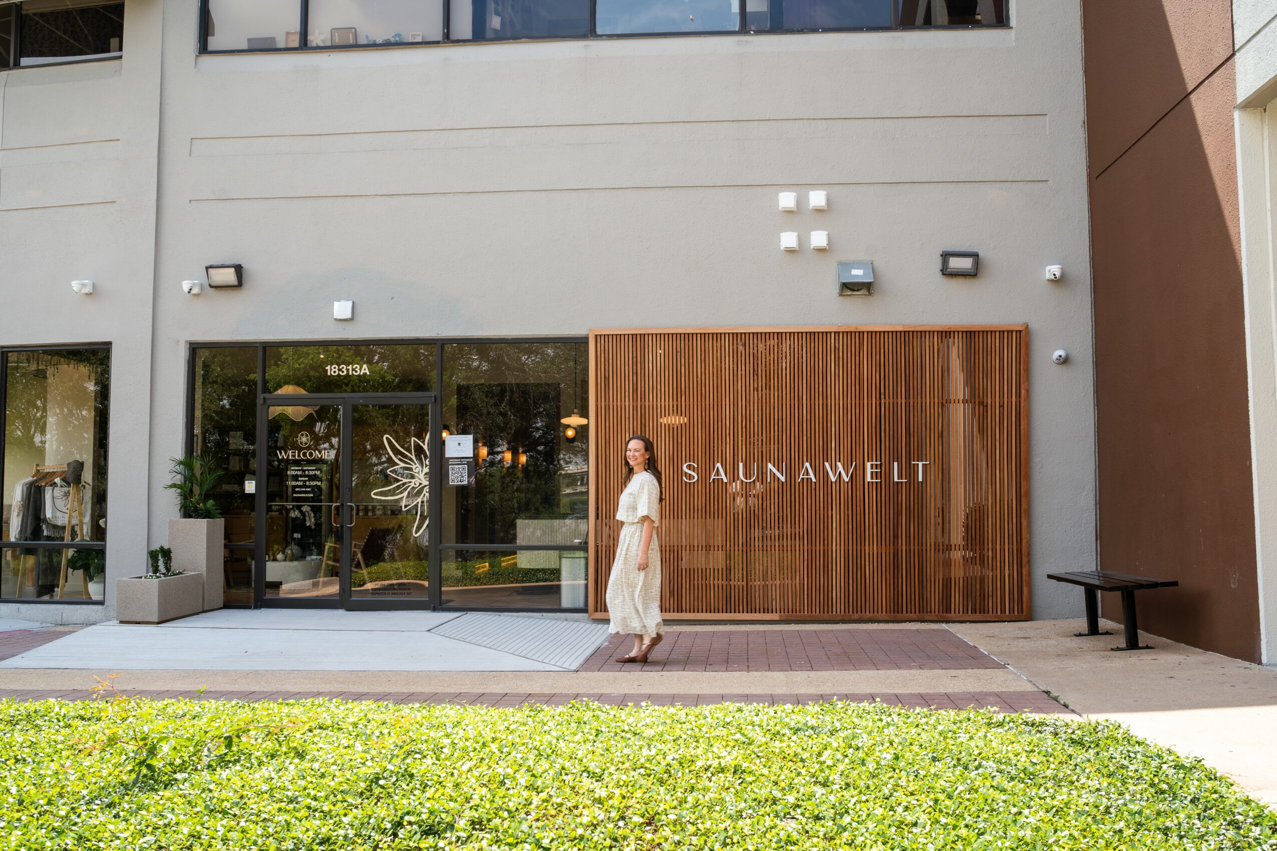 Person walking past a storefront with a wooden panel and the word 'SAUNAWEILT' on it, in front of a modern building.