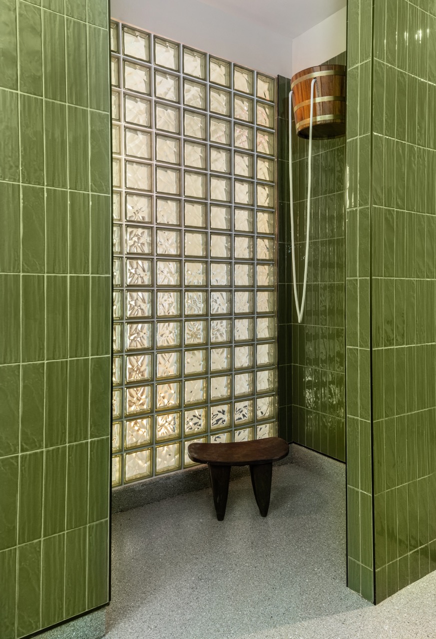 Small shower area with green tiled walls, glass block wall, wooden stool, and showerhead with wooden shelf.