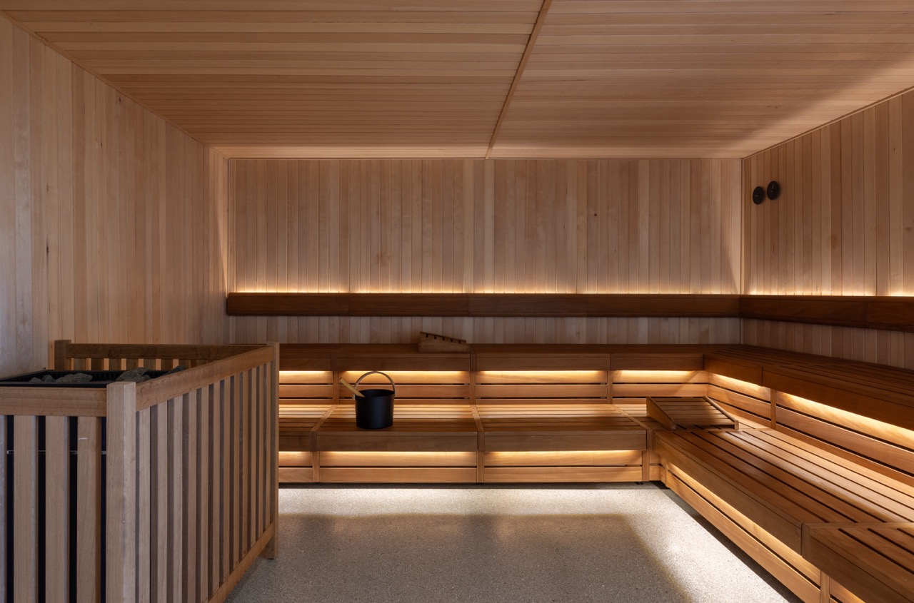 Wood-paneled sauna room with benches and a bucket, illuminated by warm lighting, and a wooden railing in the corner.