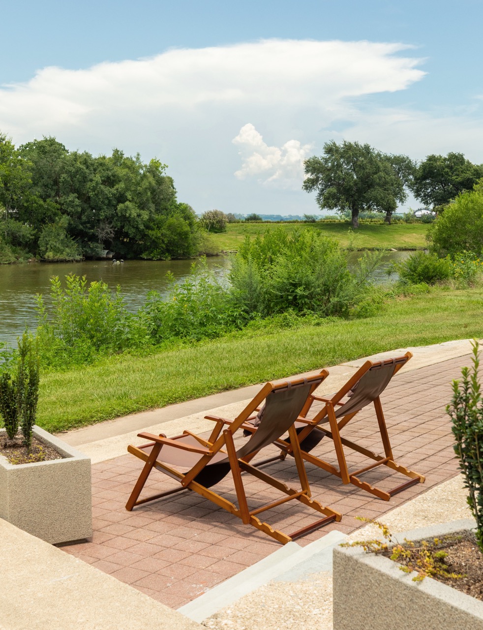 Two wooden lounge chairs on a brick patio overlooking a river and green trees under a partly cloudy sky.