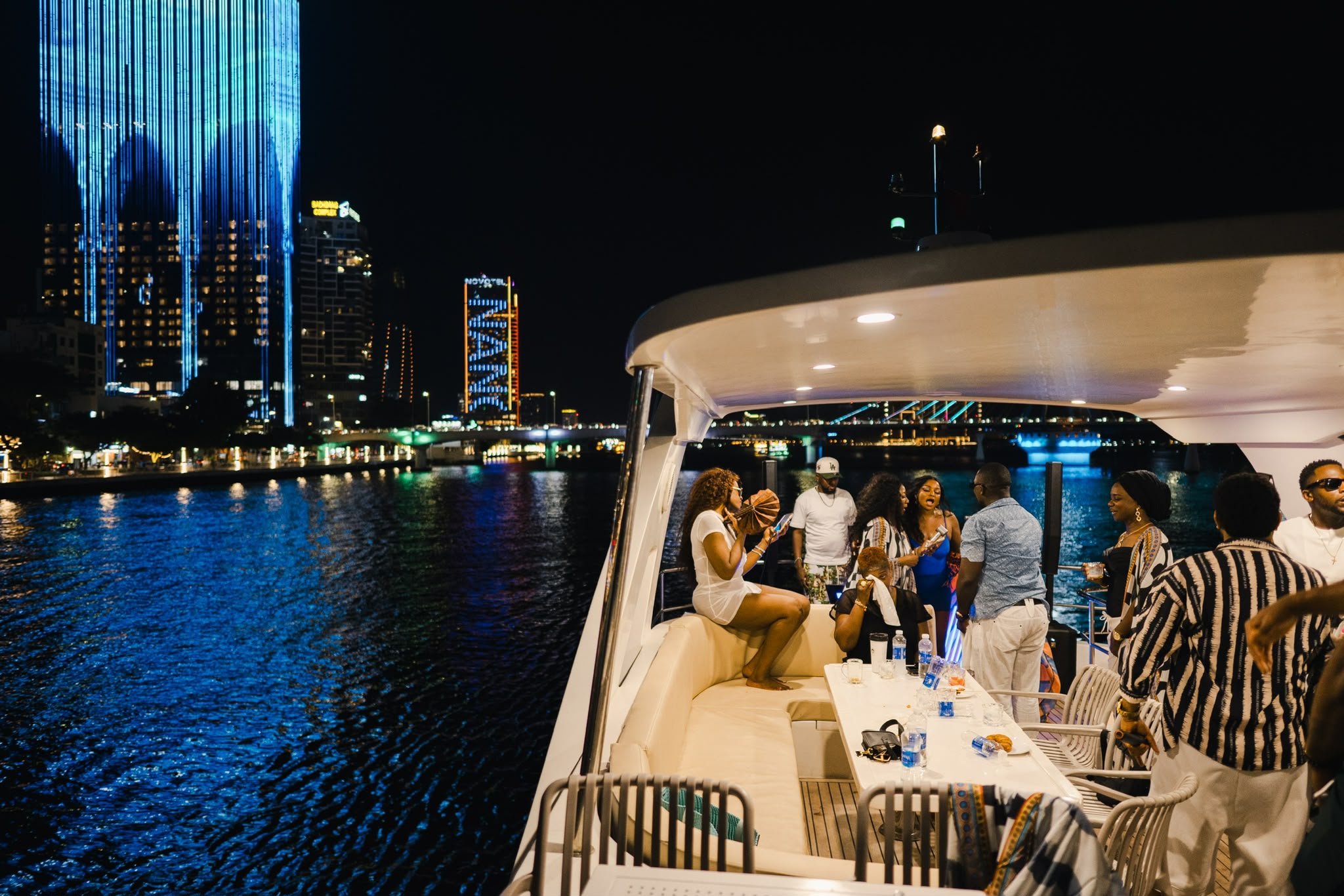 People on a boat at night with city skyline and water in the background, some standing and some seated, illuminated by city lights.