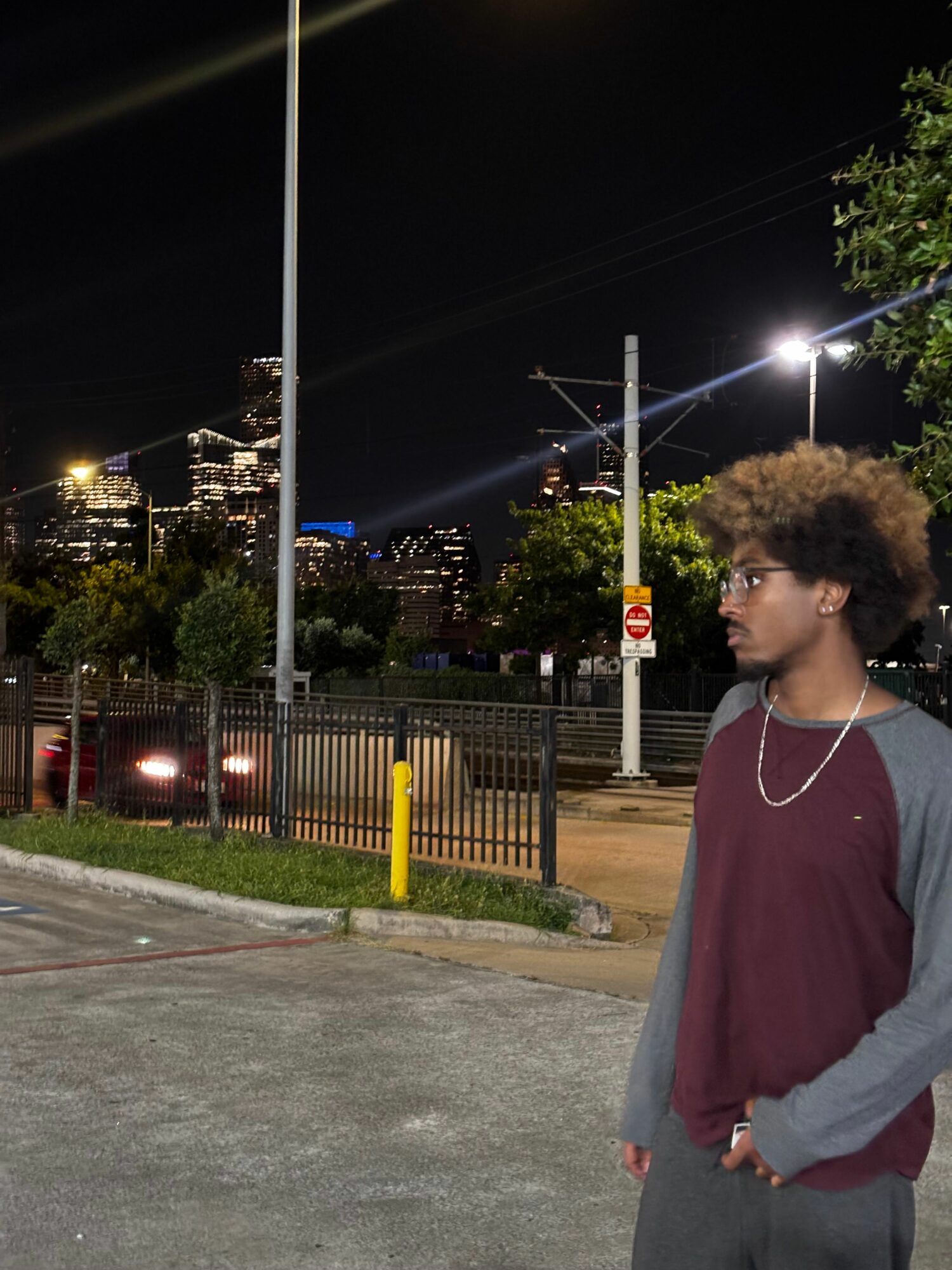 Young person with curly hair and glasses stands outdoors at night, wearing a maroon shirt and gray jacket, looking to the side.