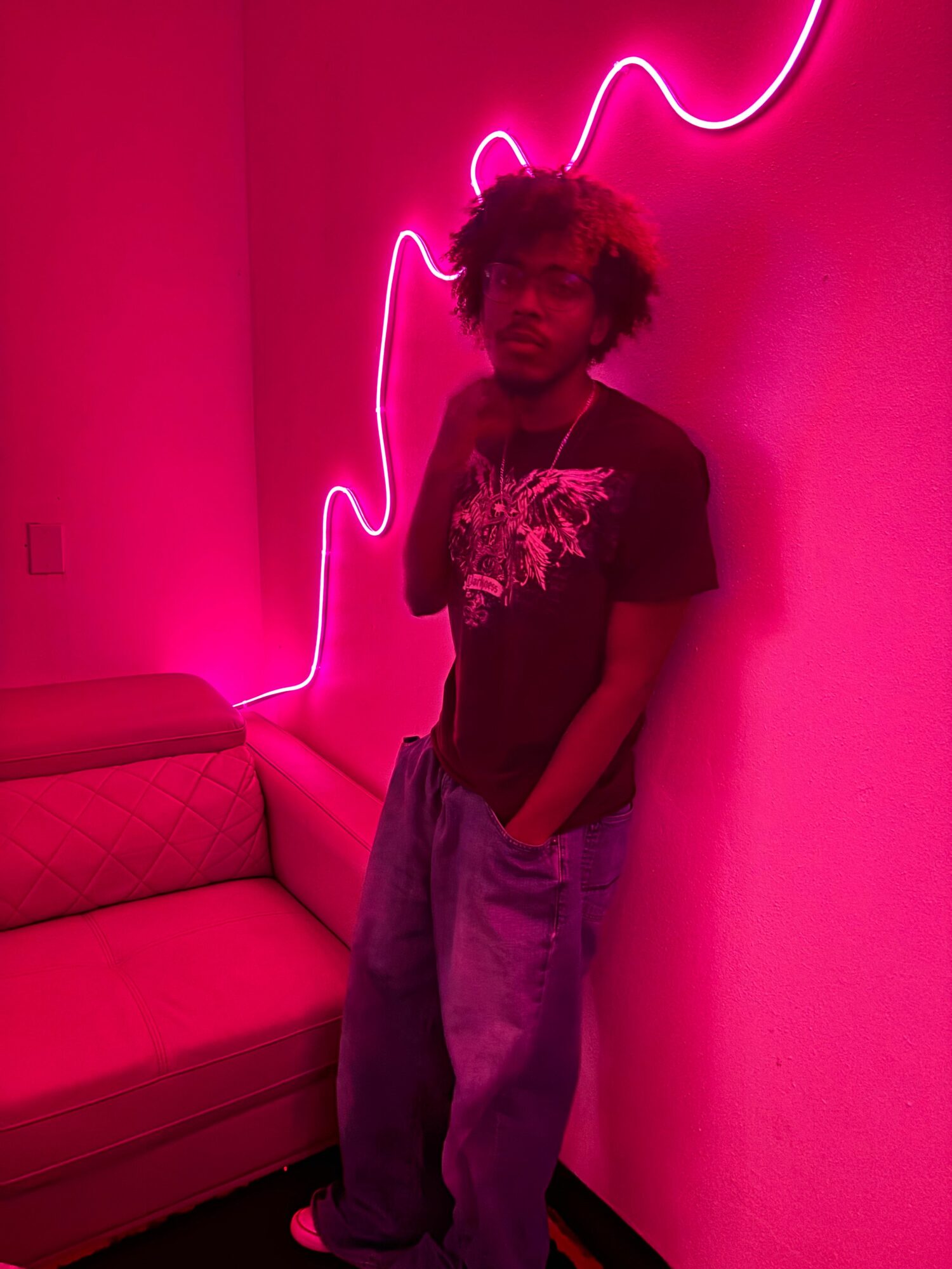 Person with curly hair standing in a pink-lit room, neon light behind them, wearing a dark shirt and jeans.