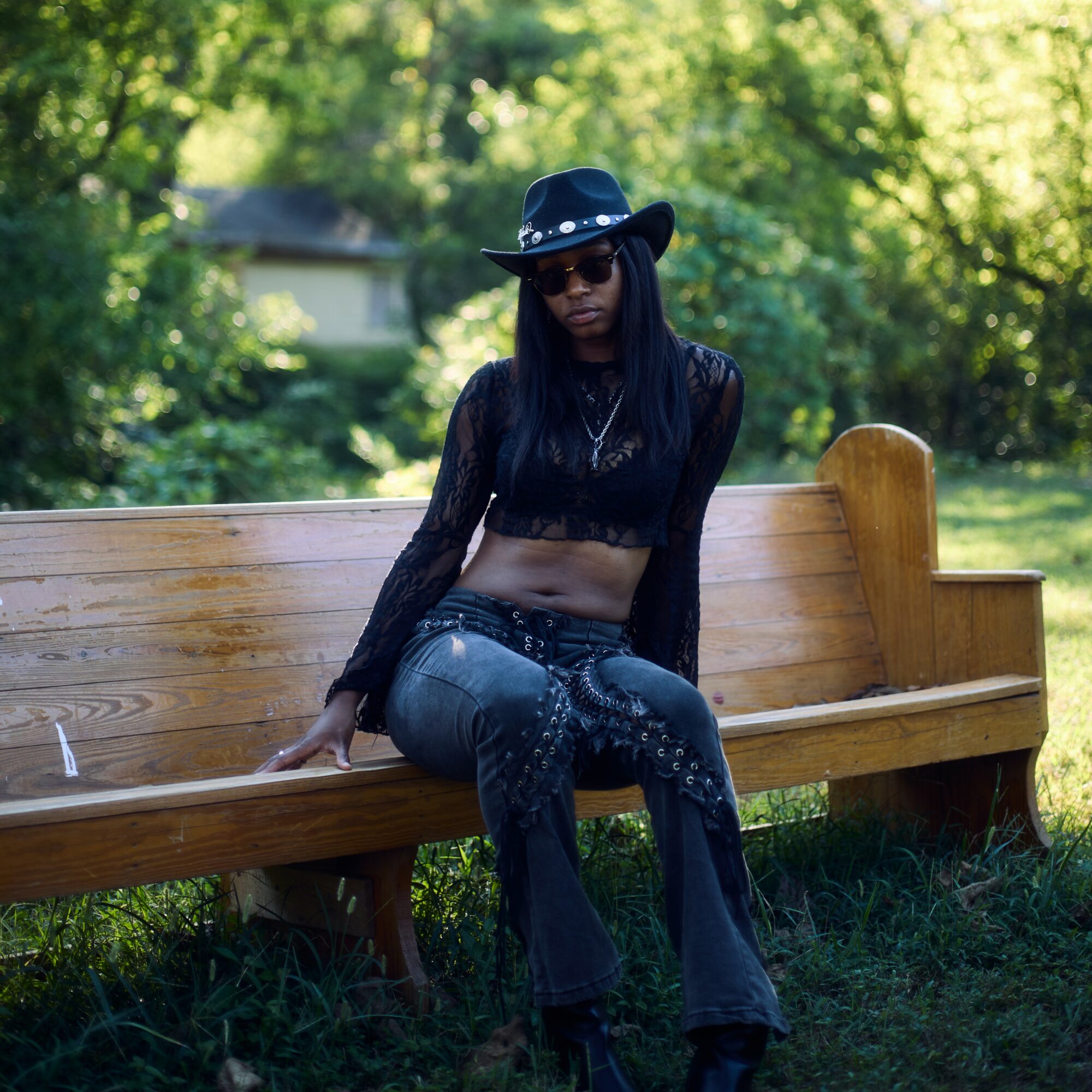 Person sitting on a wooden park bench wearing a black hat, sunglasses, a black crop top, and jeans, with trees in the background.