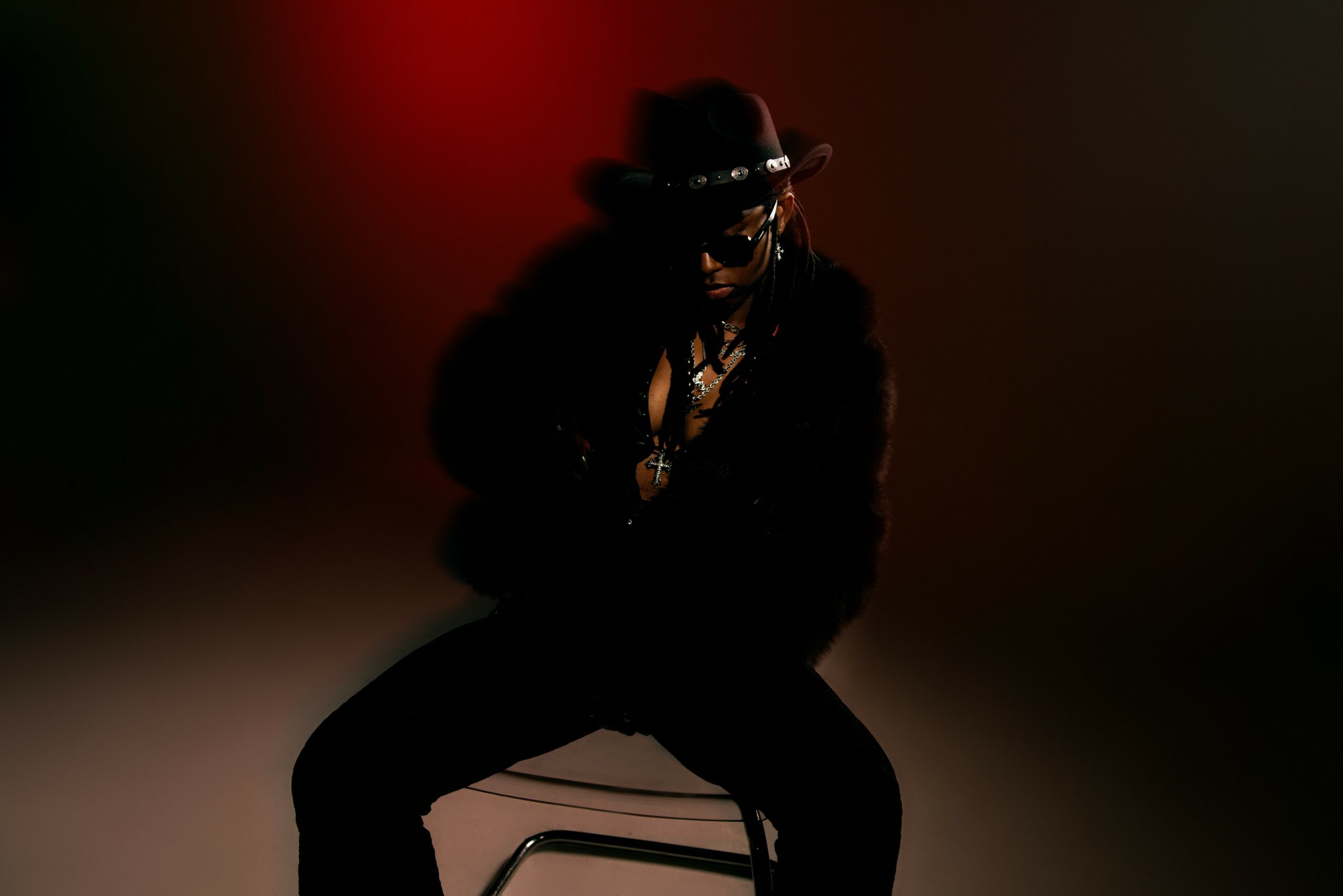 Person sitting on a chair wearing a hat, sunglasses, and dark clothing, with a dark background.