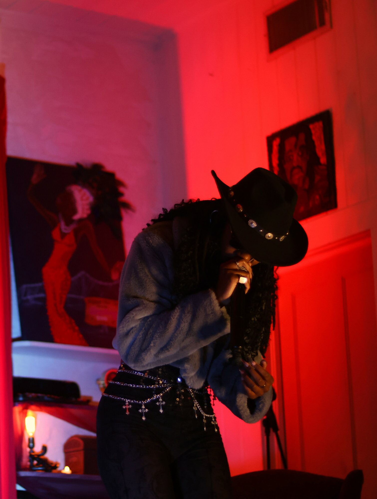 Person wearing a hat and dark clothing, holding a microphone, in a room with red lighting and artwork on the wall.