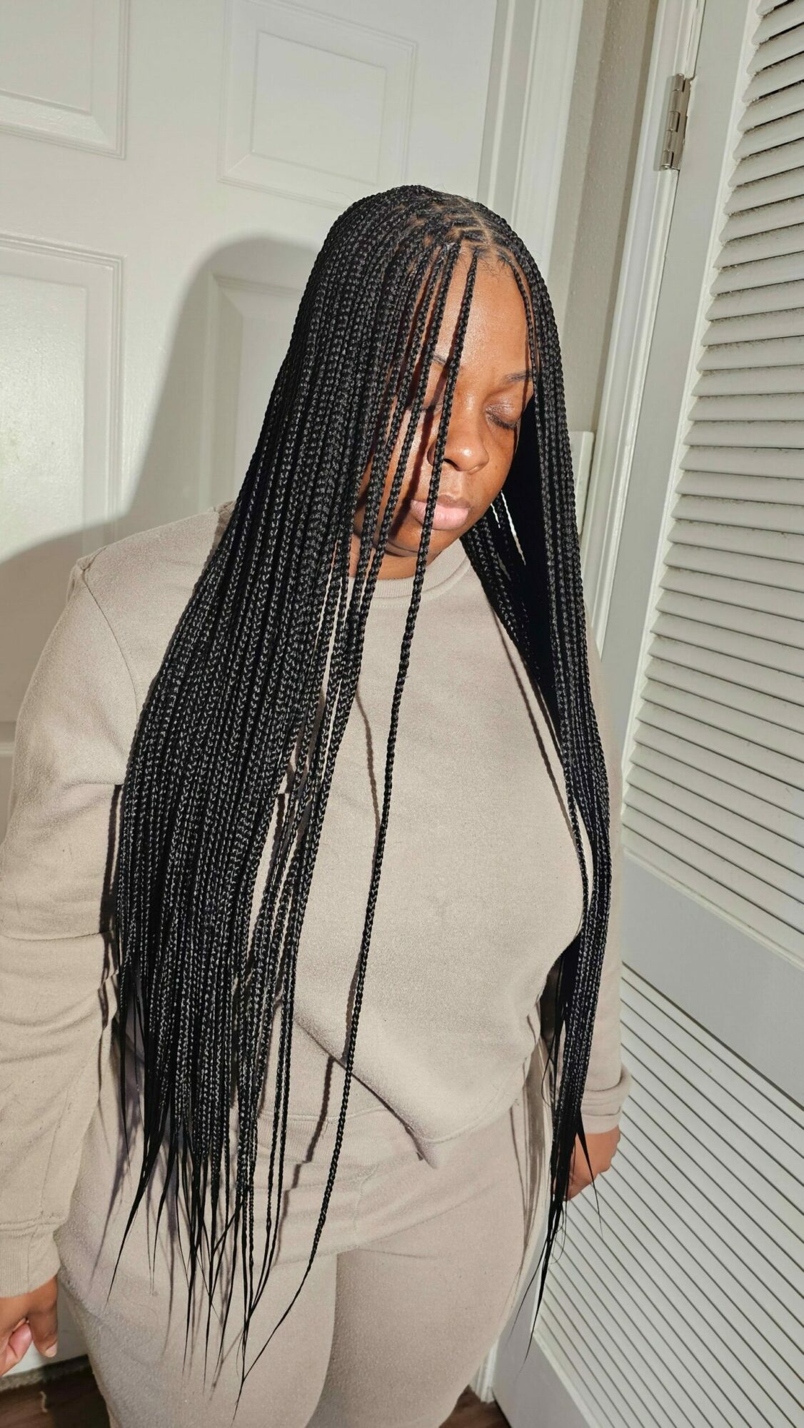 Person with long braided hair wearing a beige top, standing indoors against a white door and wall.