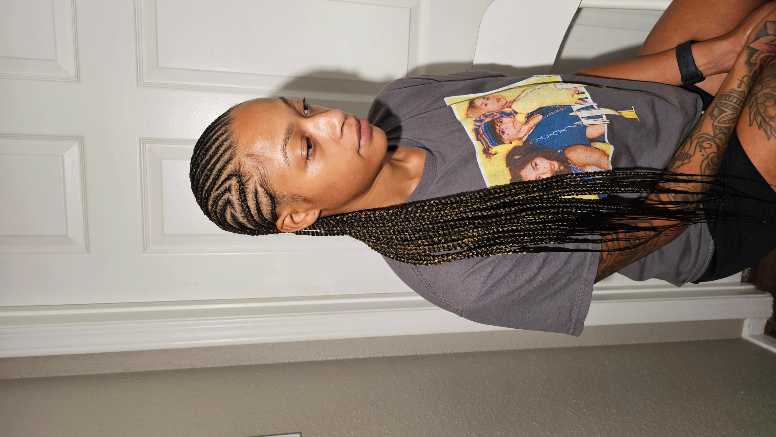 Woman with long braided hair, wearing a graphic t-shirt, sitting against a white door, looking to the side.