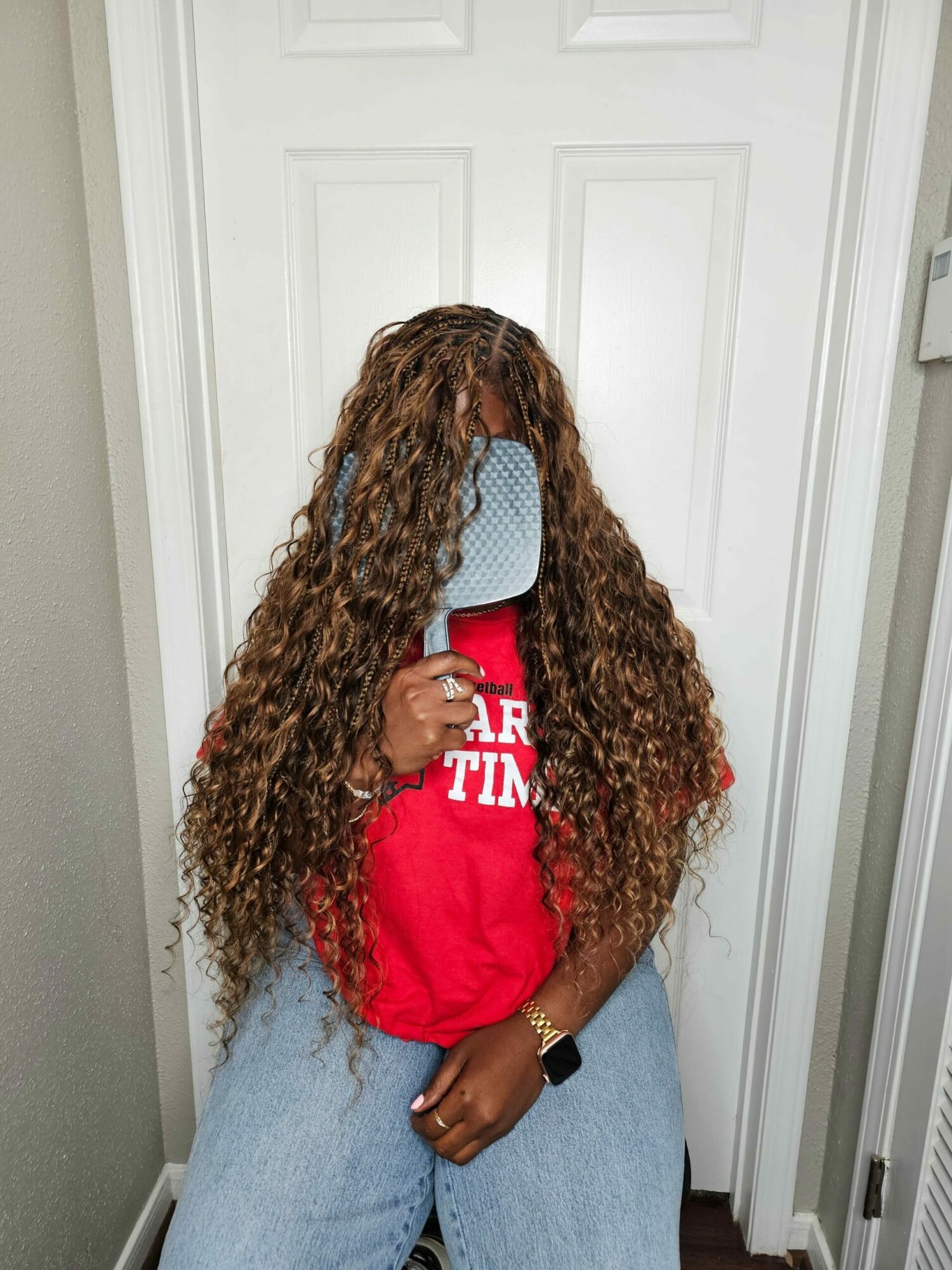 Person with long curly hair wearing a red shirt and jeans, standing indoors near a white door.