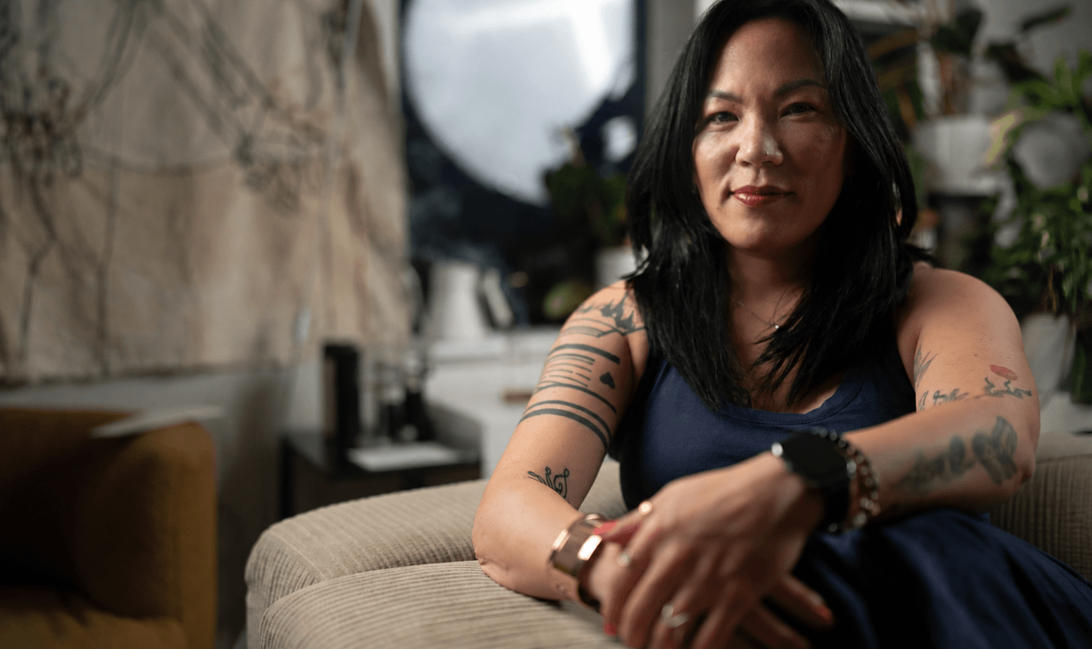 Woman with black hair and tattoos sitting on a beige couch in a room with plants and a mirror.