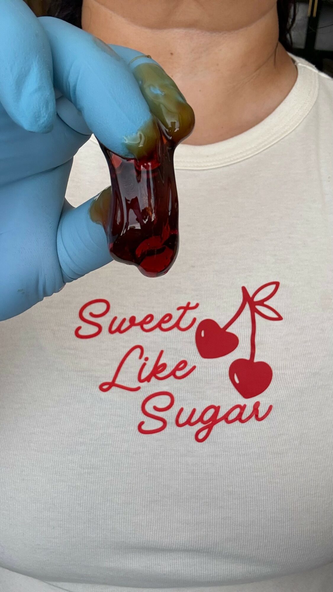 Person wearing a white shirt with red text and cherries, holding a green and red gummy candy, with a blue glove.