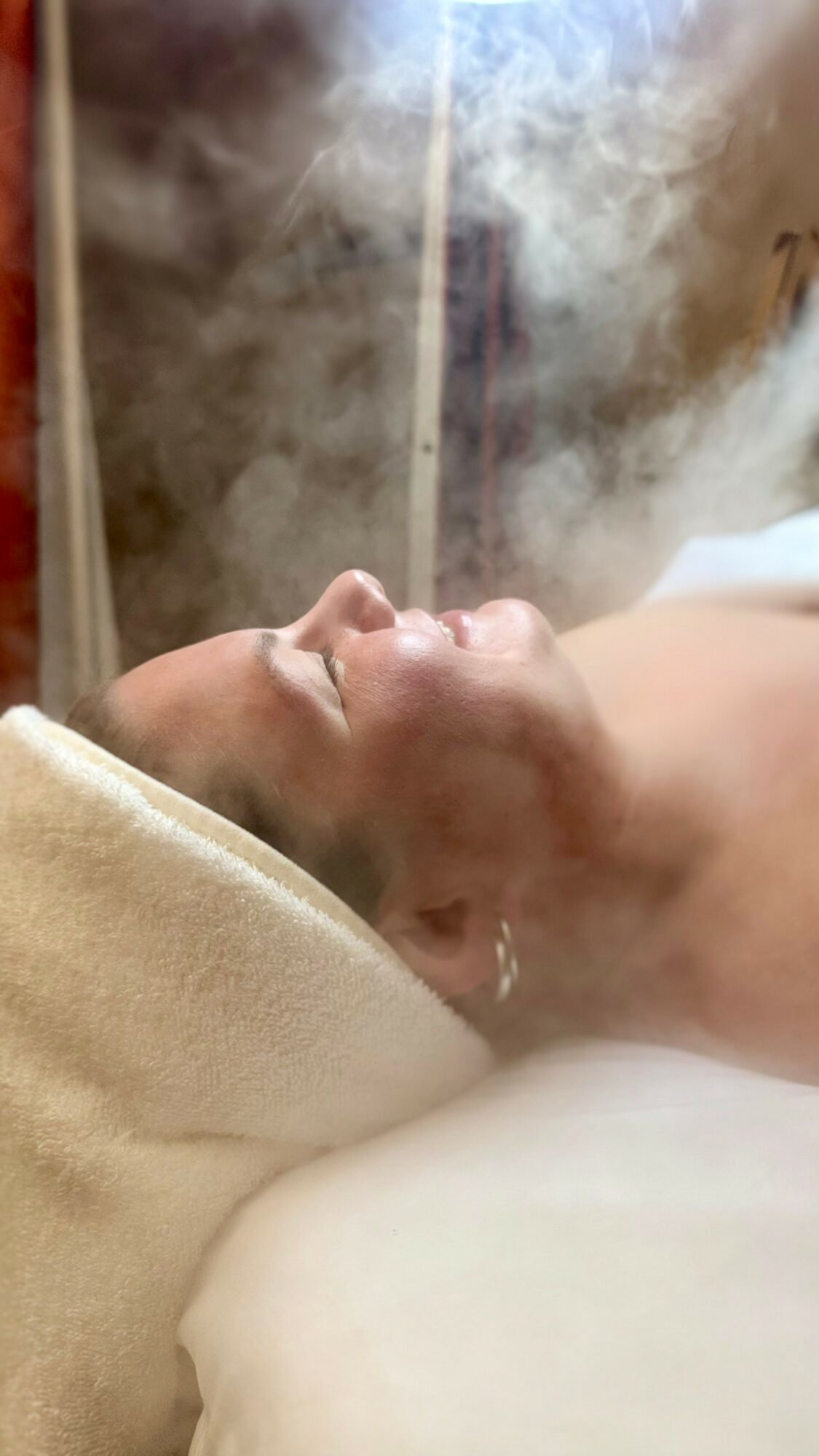 Person lying down with head tilted back, surrounded by steam or smoke, in a spa or sauna setting.