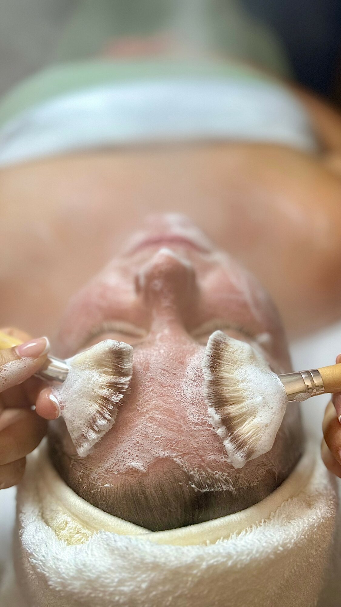 Person receiving facial treatment with brushes on cheeks, lying down with head wrapped in towel, eyes closed.