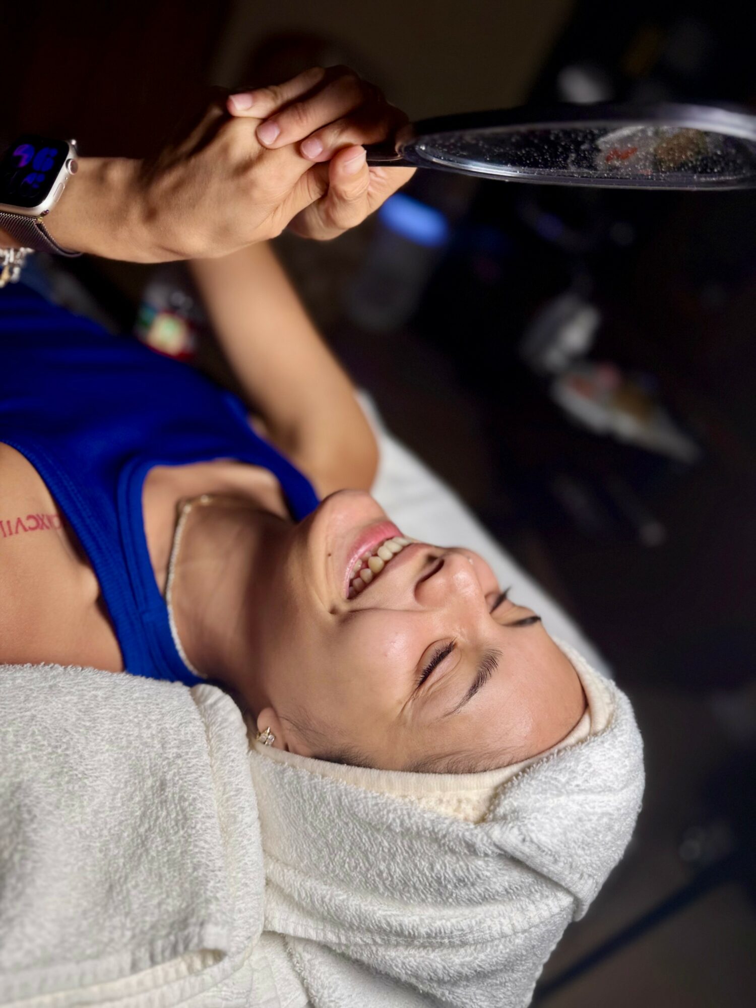 Woman lying down with a towel wrapped around her head, smiling, holding a glass, in a spa or wellness setting.