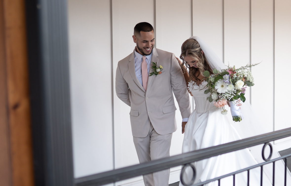A bride and groom walk together, smiling, with the bride holding a bouquet of flowers, against a plain wall background.