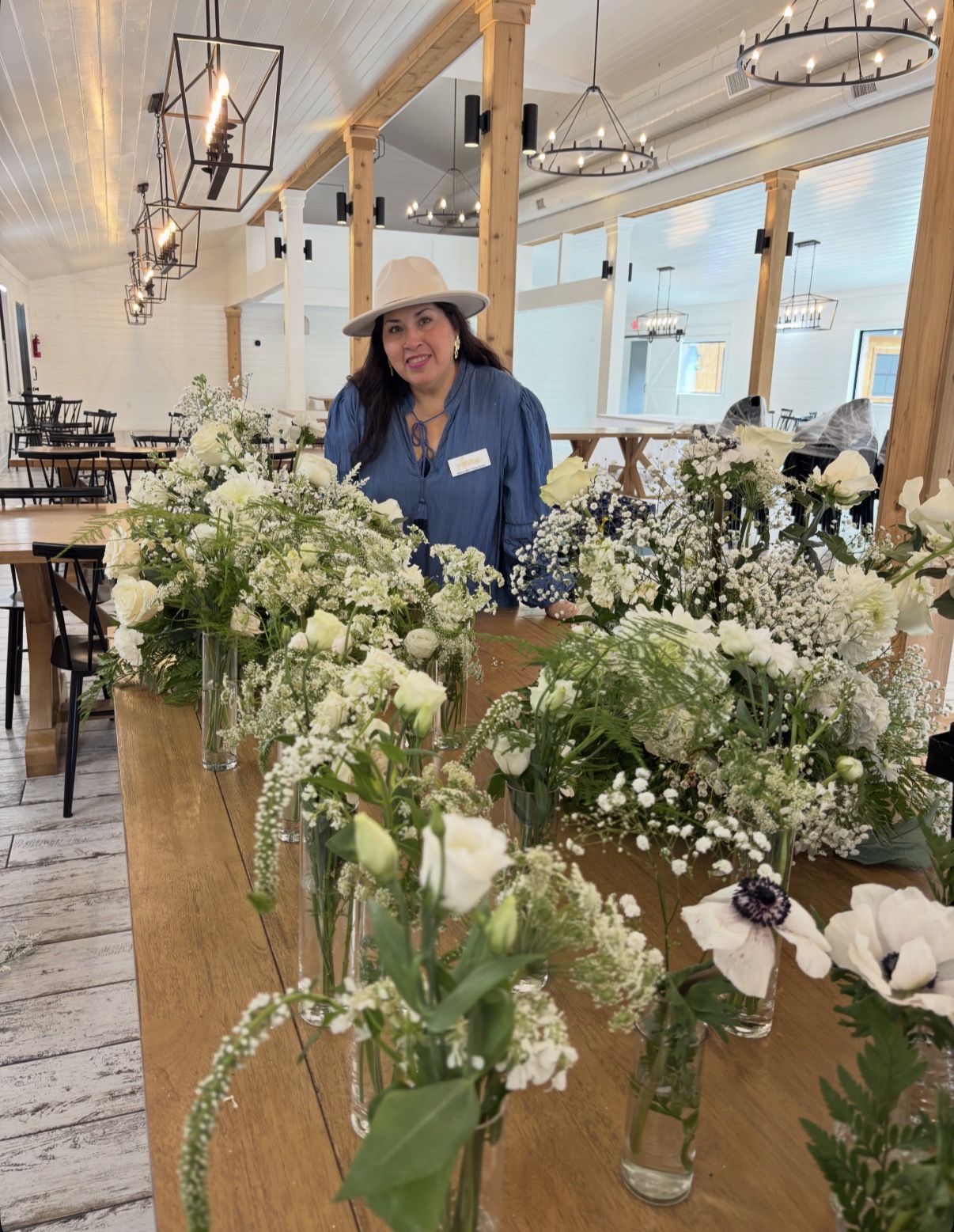 Woman wearing a hat and blue jacket standing behind a table with white and purple flowers in a bright, modern indoor space.