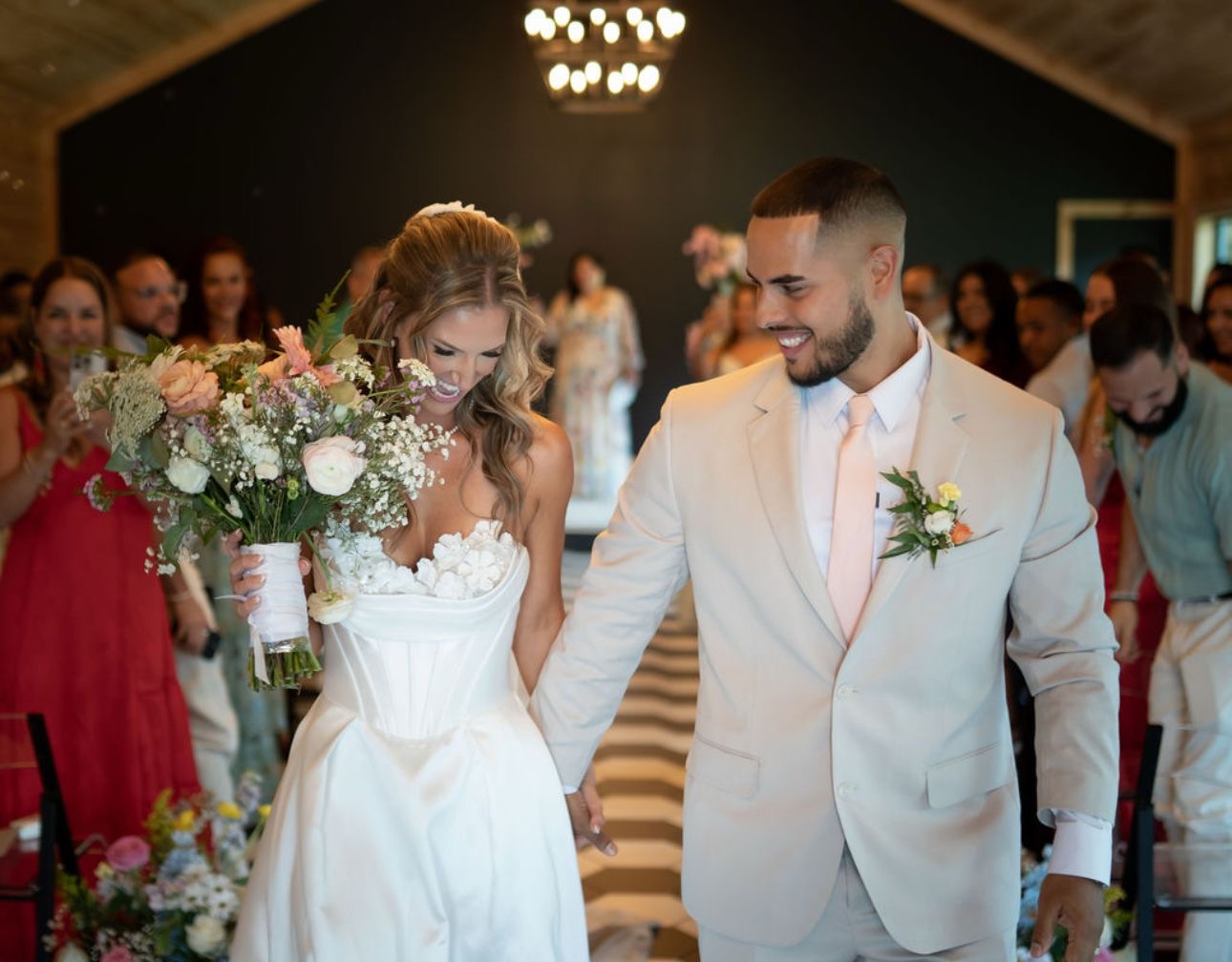 Bride and groom holding hands and smiling at each other during wedding ceremony, guests in background, indoor setting.