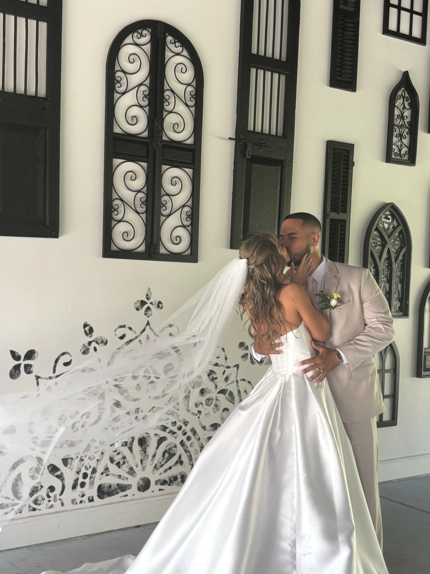 Bride and groom embrace, bride in a white wedding gown with veil, standing against a wall with decorative black window frames.