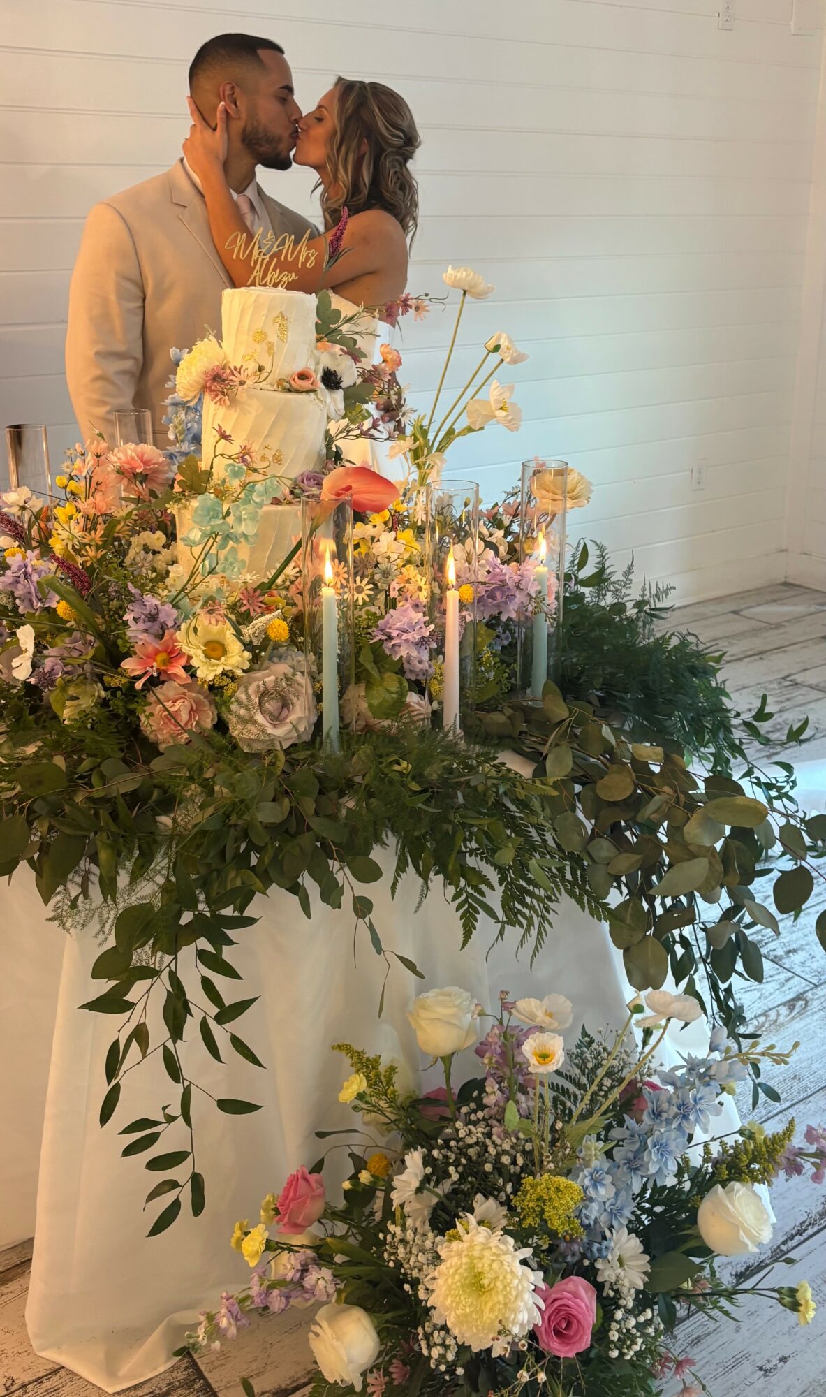 Couple kissing behind a decorated table with flowers and candles at a celebration, possibly a wedding, indoors.