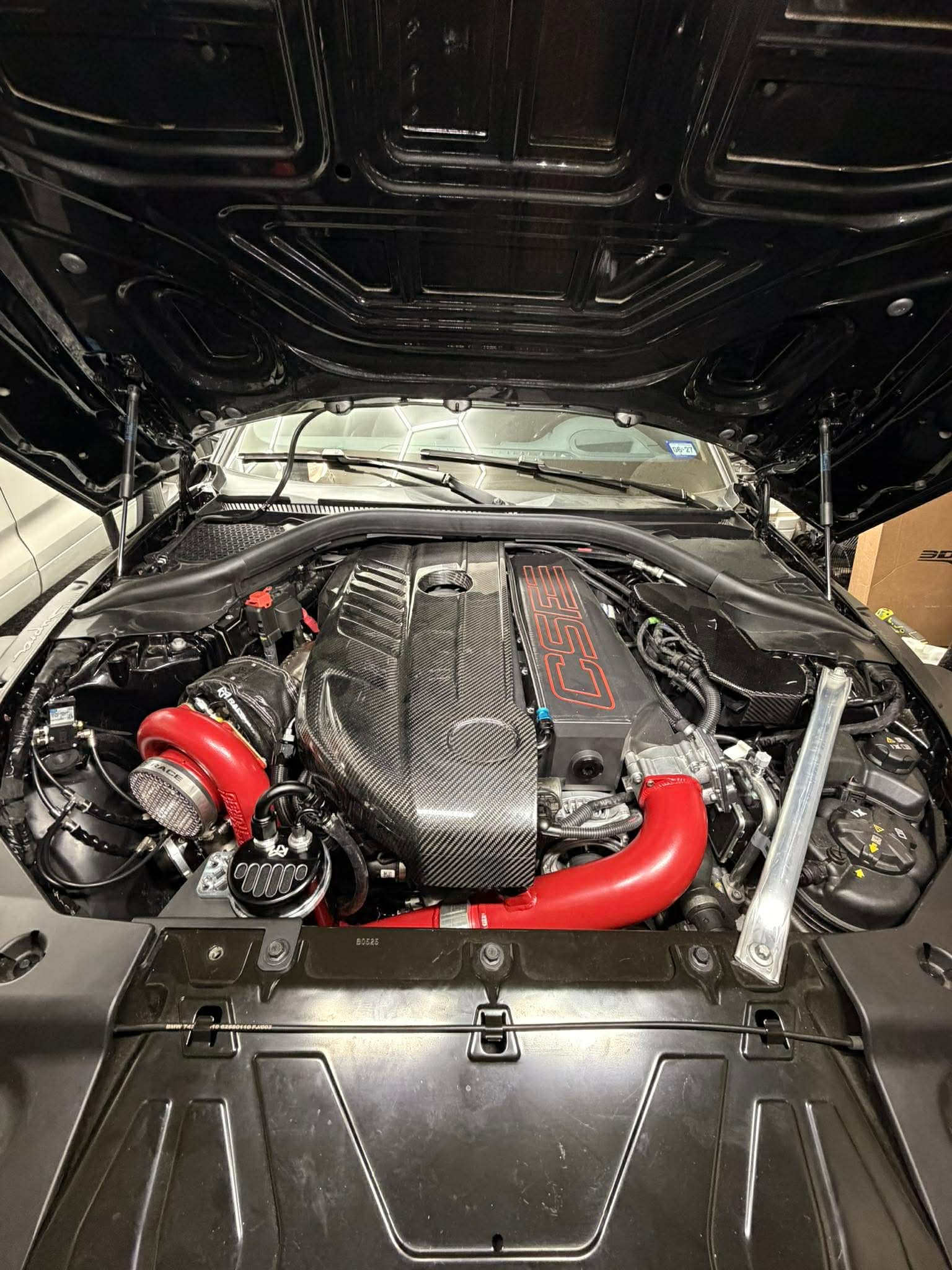 Car engine with red intake pipe, black engine cover, and various components visible under open hood.