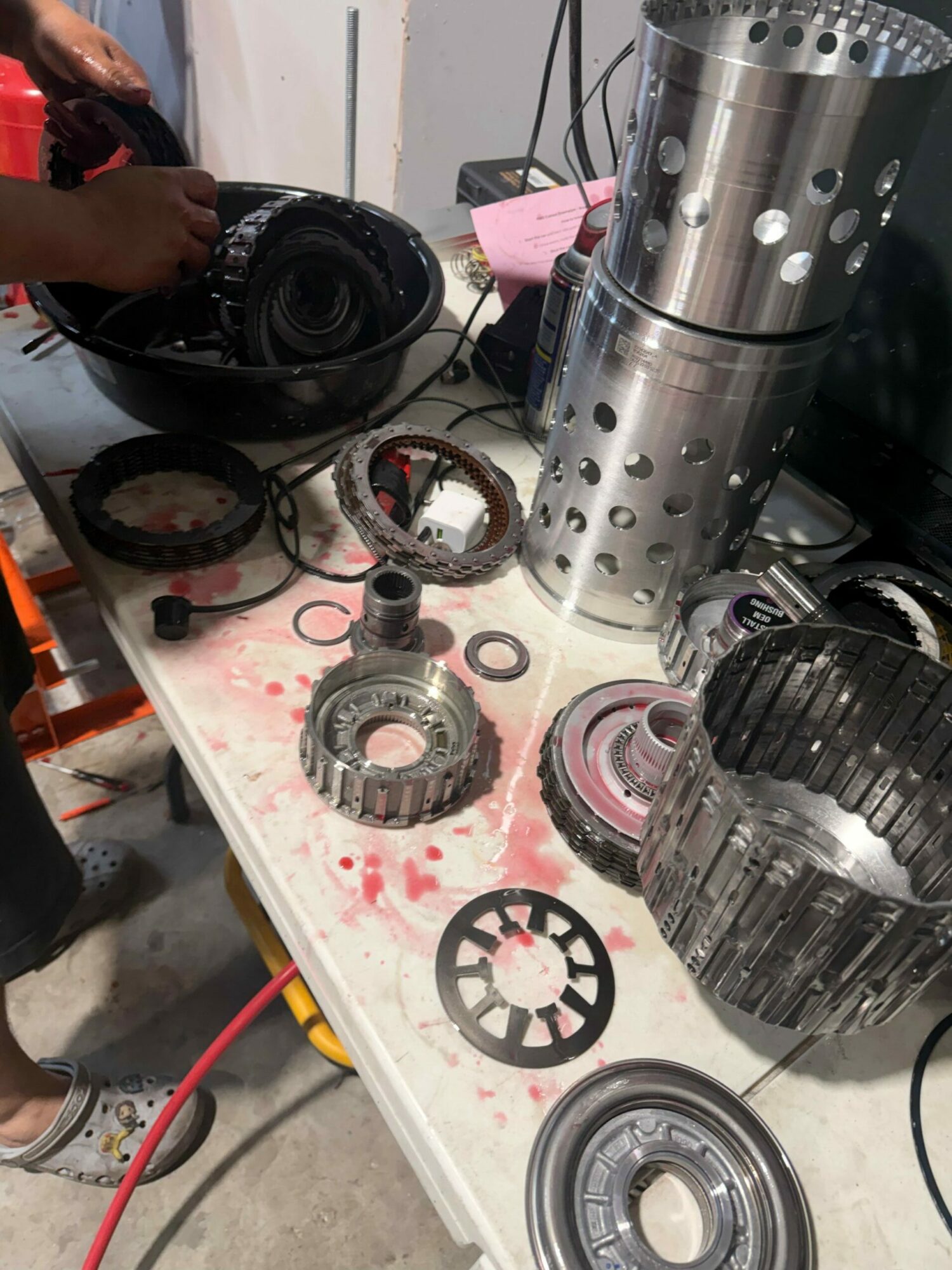 Disassembled mechanical parts, gears, and tools on a white work surface with some red stains, viewed from above.
