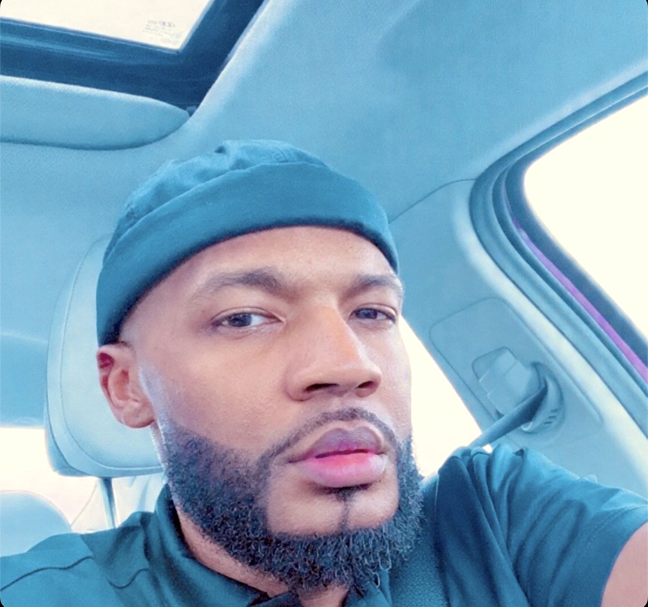 Man with beard wearing a beanie inside a car, looking at the camera, with a light-colored interior and a sunroof.