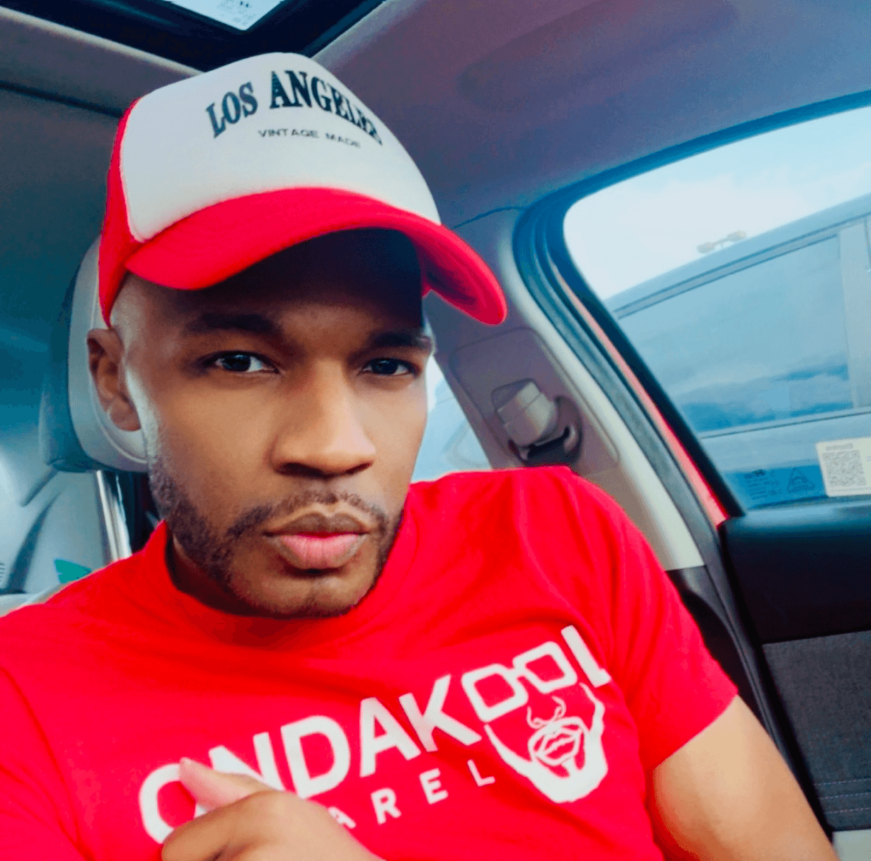 Young man wearing a red and white Los Angeles cap and red shirt inside a car.