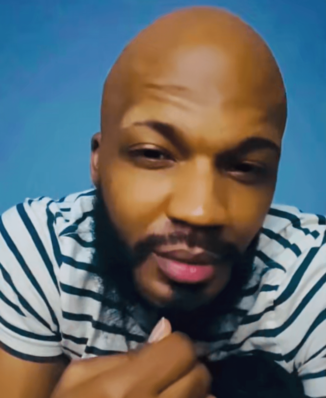 Close-up of a person with a bald head and beard, wearing a striped shirt, against a blue background.