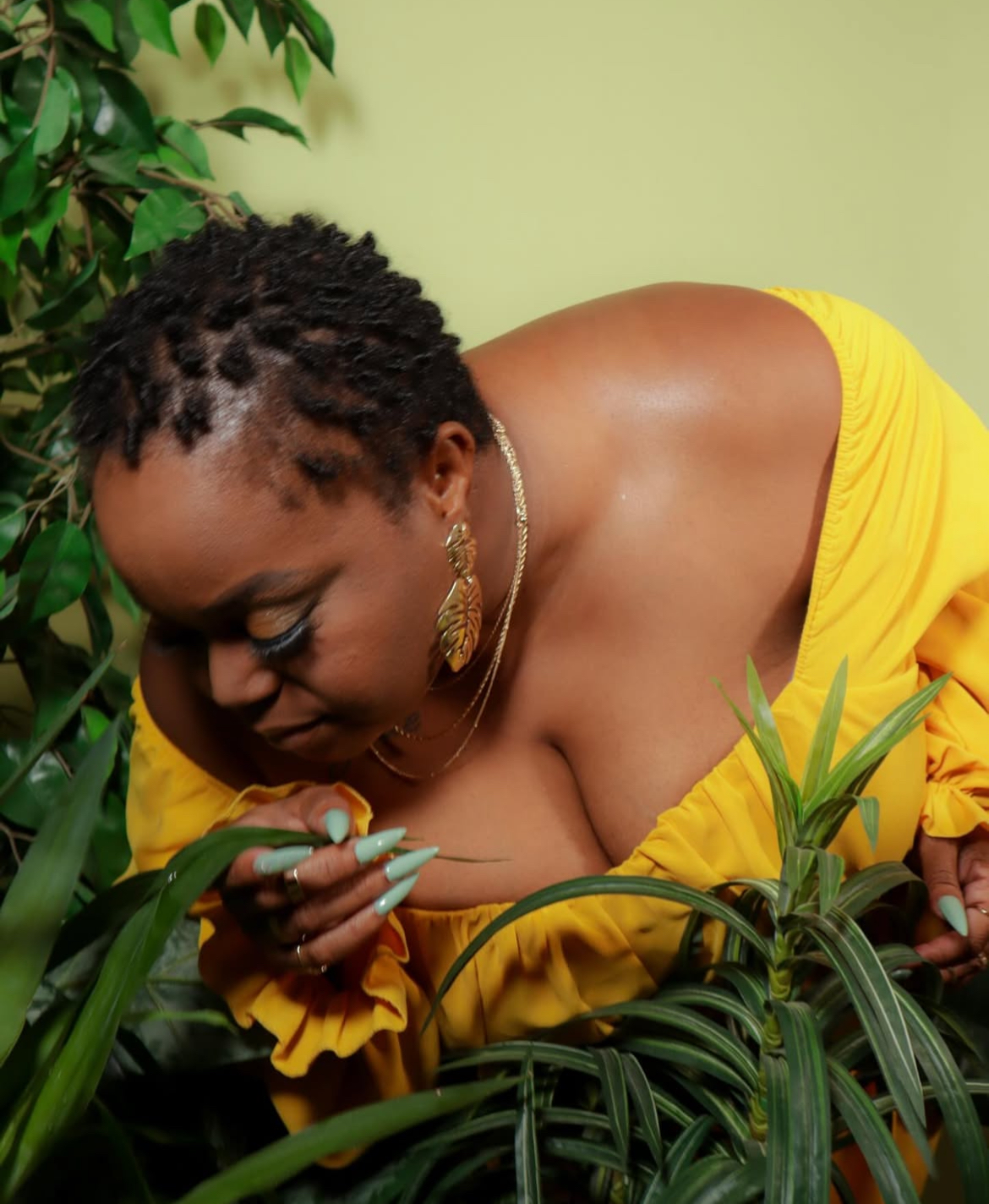 Woman with short curly hair wearing yellow dress, jewelry, leaning among green plants, looking down at leaves.