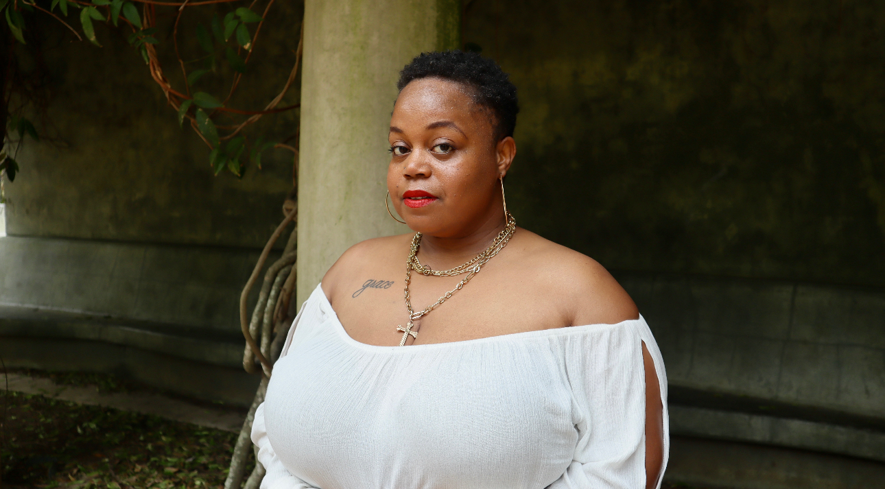 Woman with short hair wearing an off-shoulder white top outdoors, standing near a tree and a wooden fence.