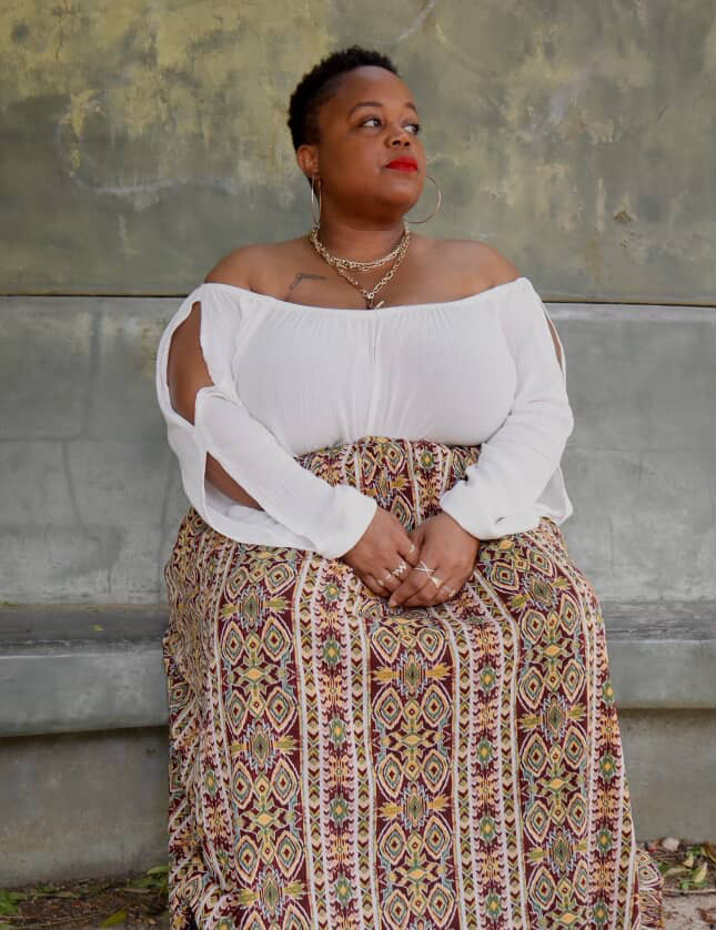 Woman sitting against a concrete wall, wearing a white off-shoulder top and patterned skirt, looking to the side.