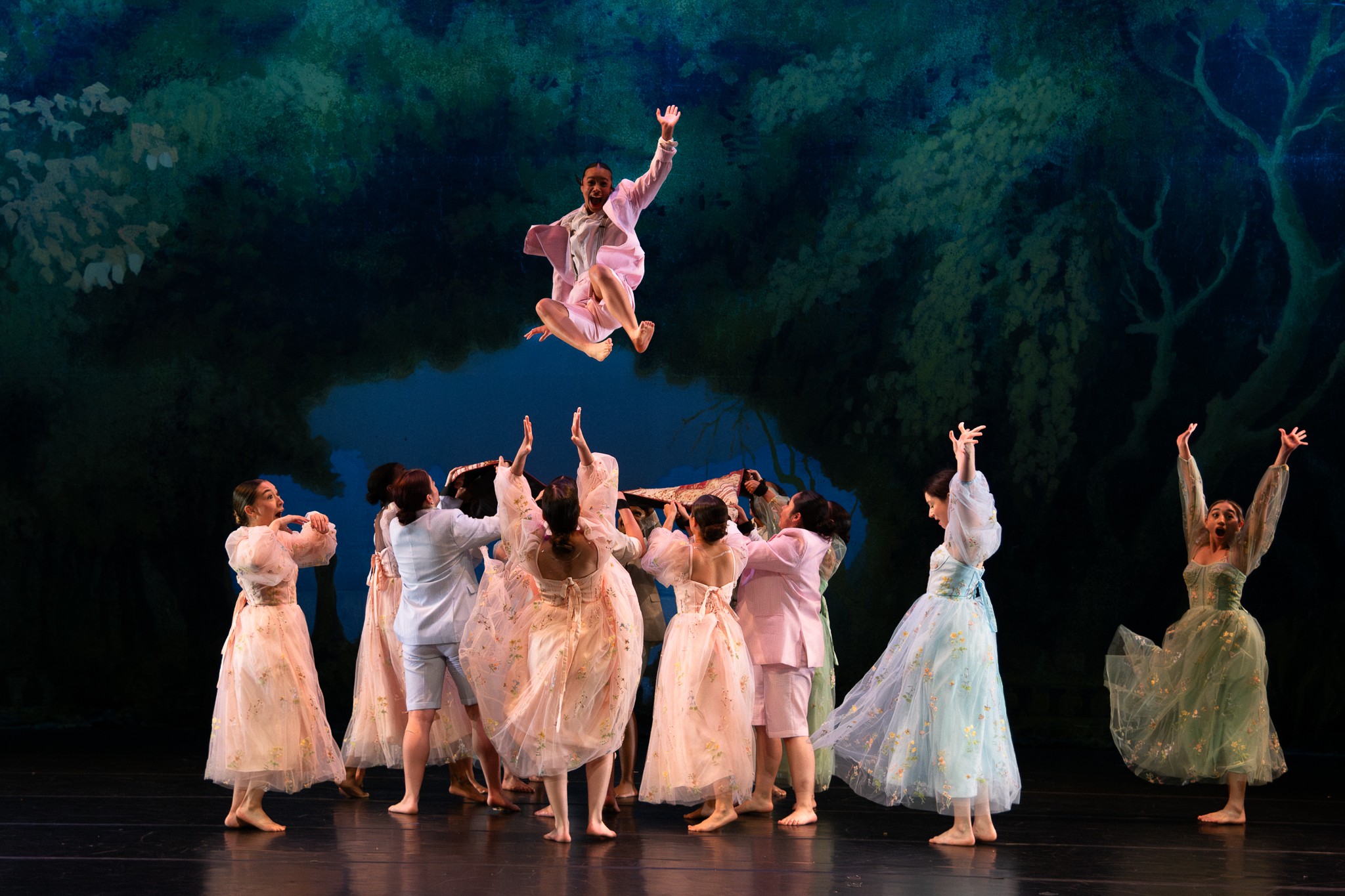 A group of dancers in pastel costumes perform on stage with a forest backdrop, one dancer is airborne above others.