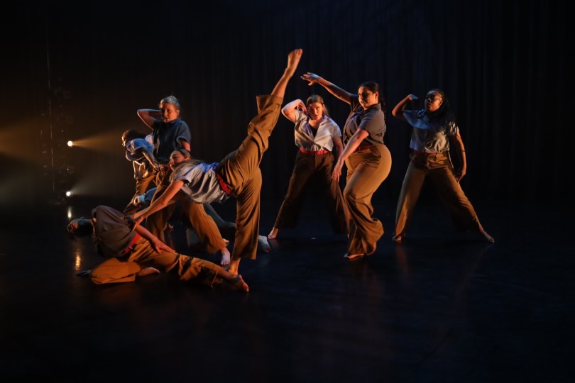Group of dancers performing on stage with dramatic lighting, some in dynamic poses and others standing in the background.