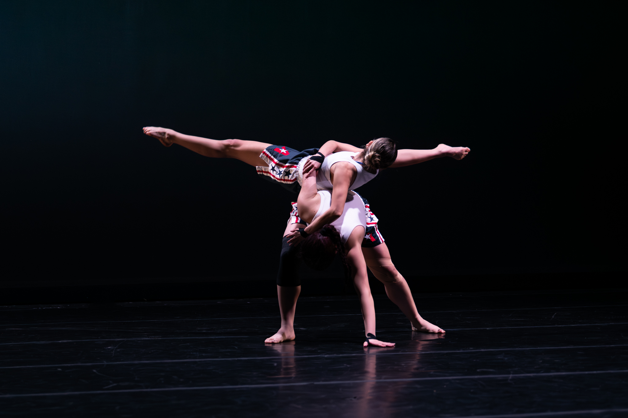Two dancers perform on stage, one supporting the other in a balancing pose with arms extended, against a dark background.