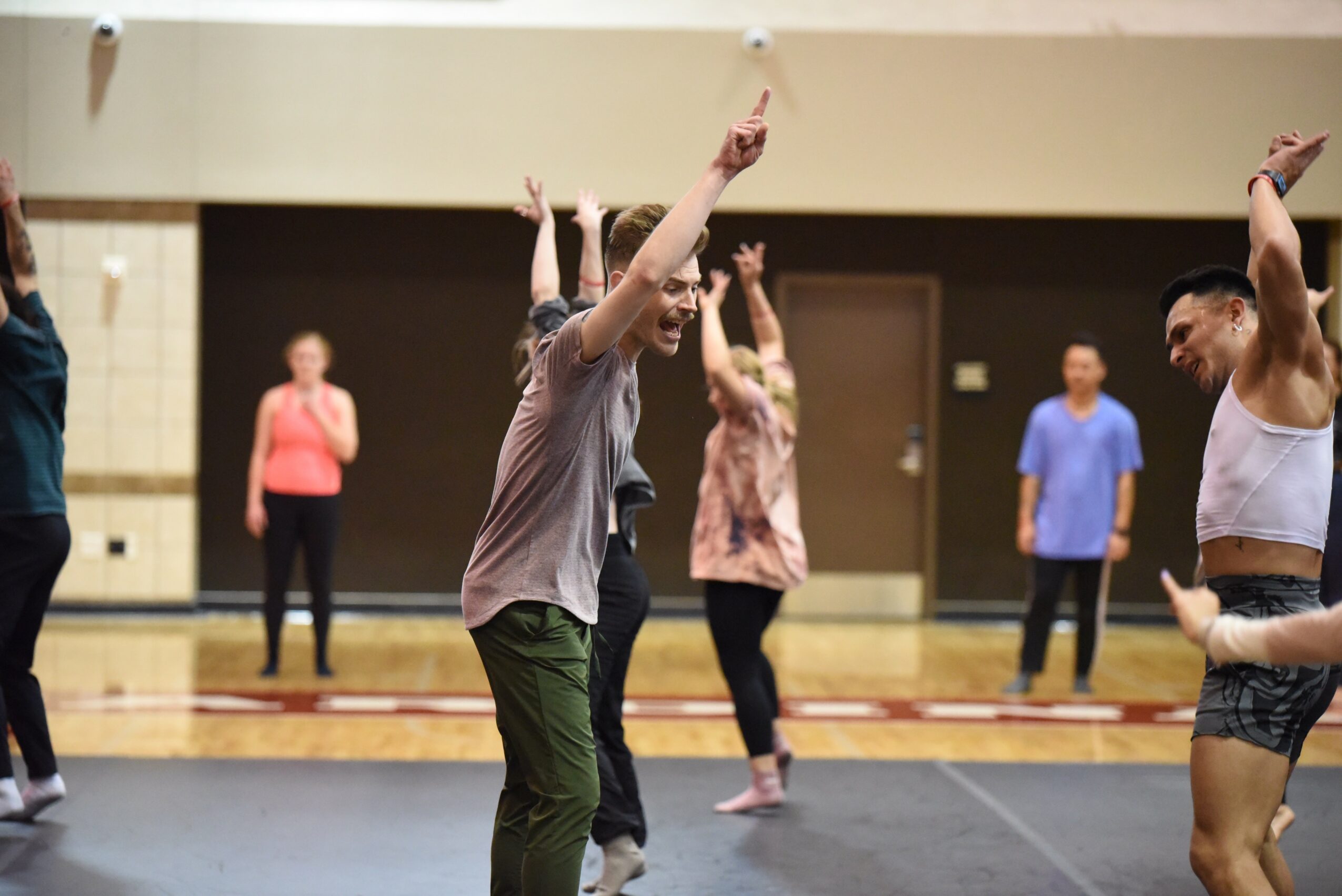 Group of people dancing in a gymnasium, some with arms raised, others standing and watching, with a mirror and wooden floor.
