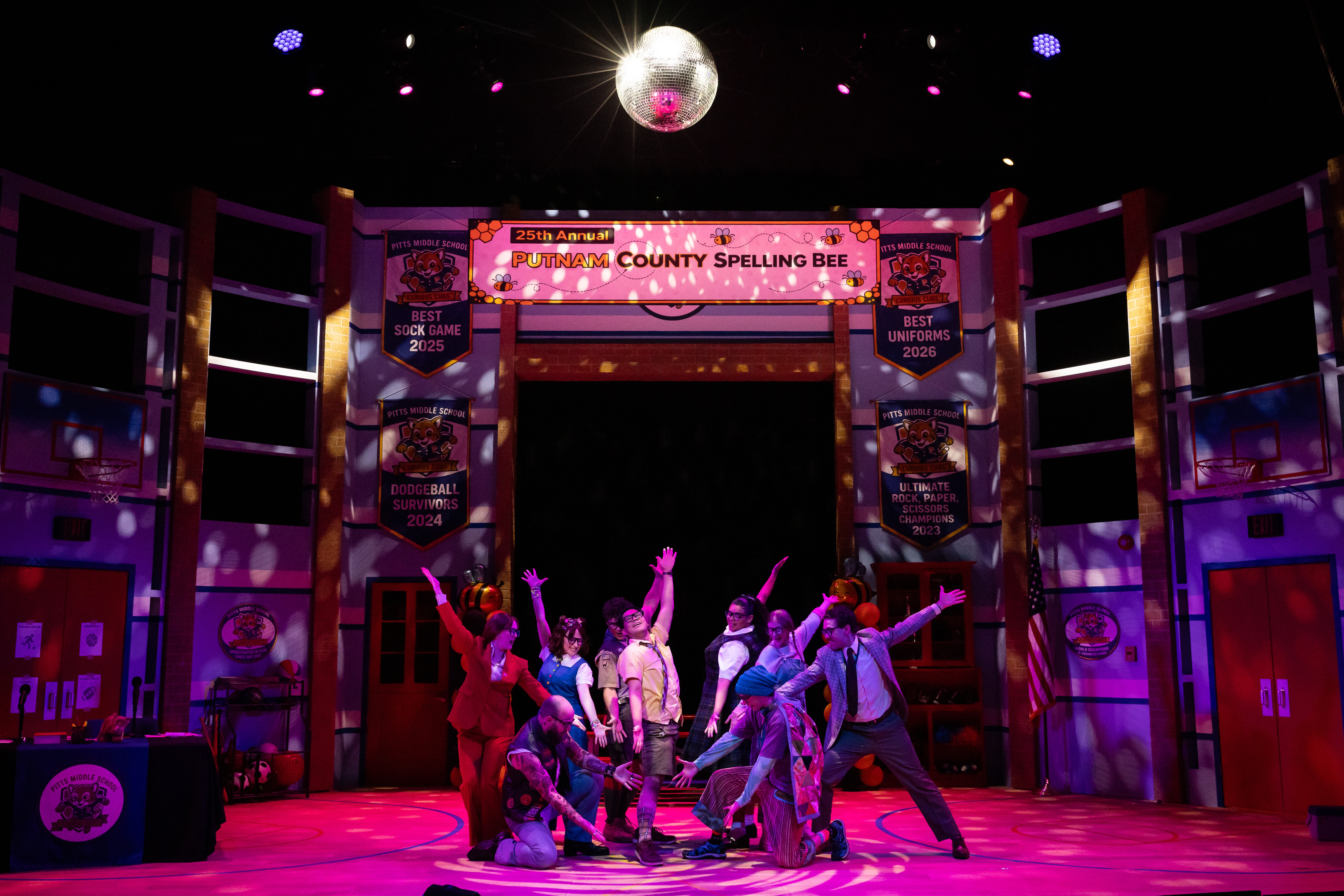 Group of children performing on stage during a spelling bee event, with colorful lighting and a disco ball overhead.