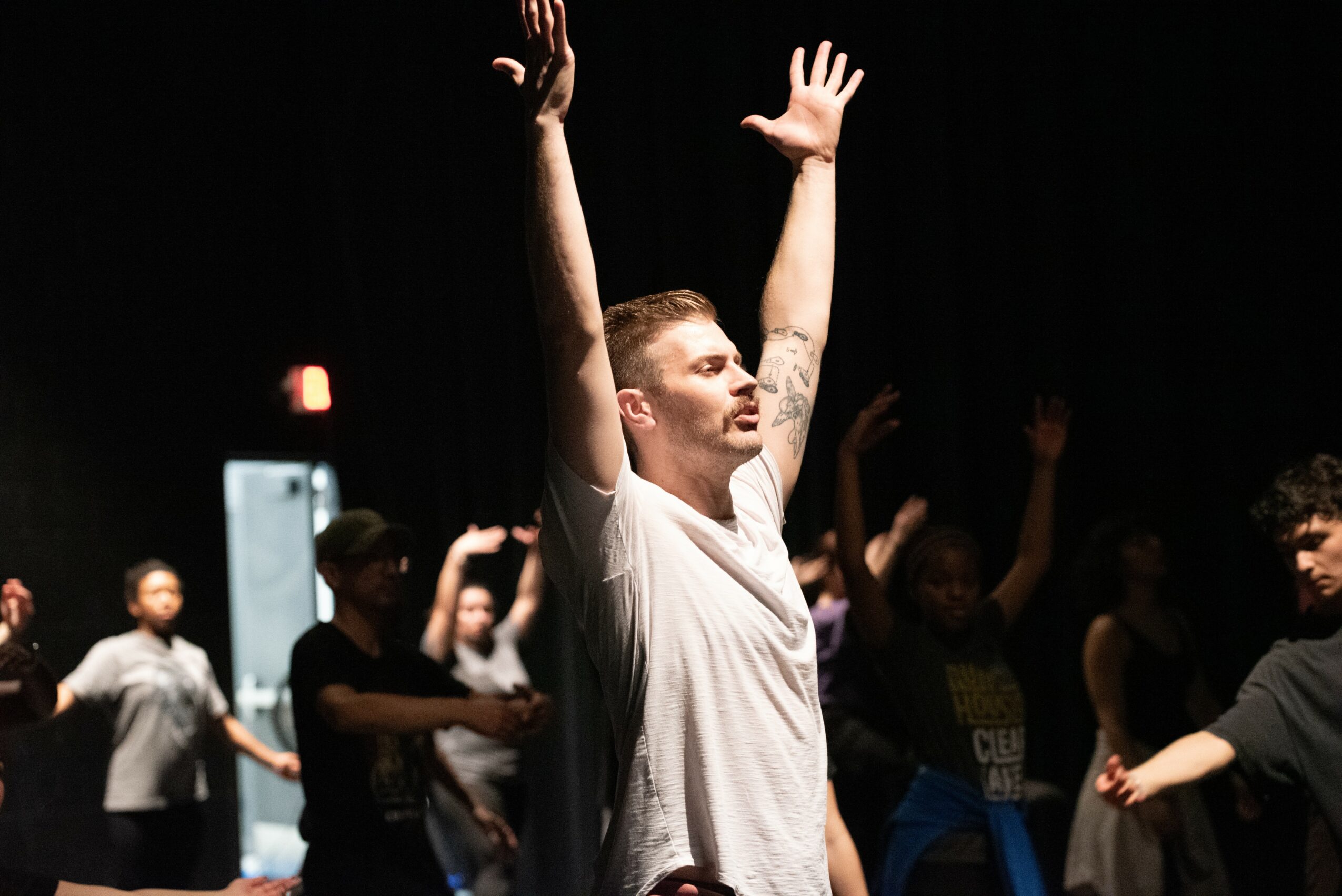 Man with arms raised in a dark room with other people in the background, some also raising their hands.